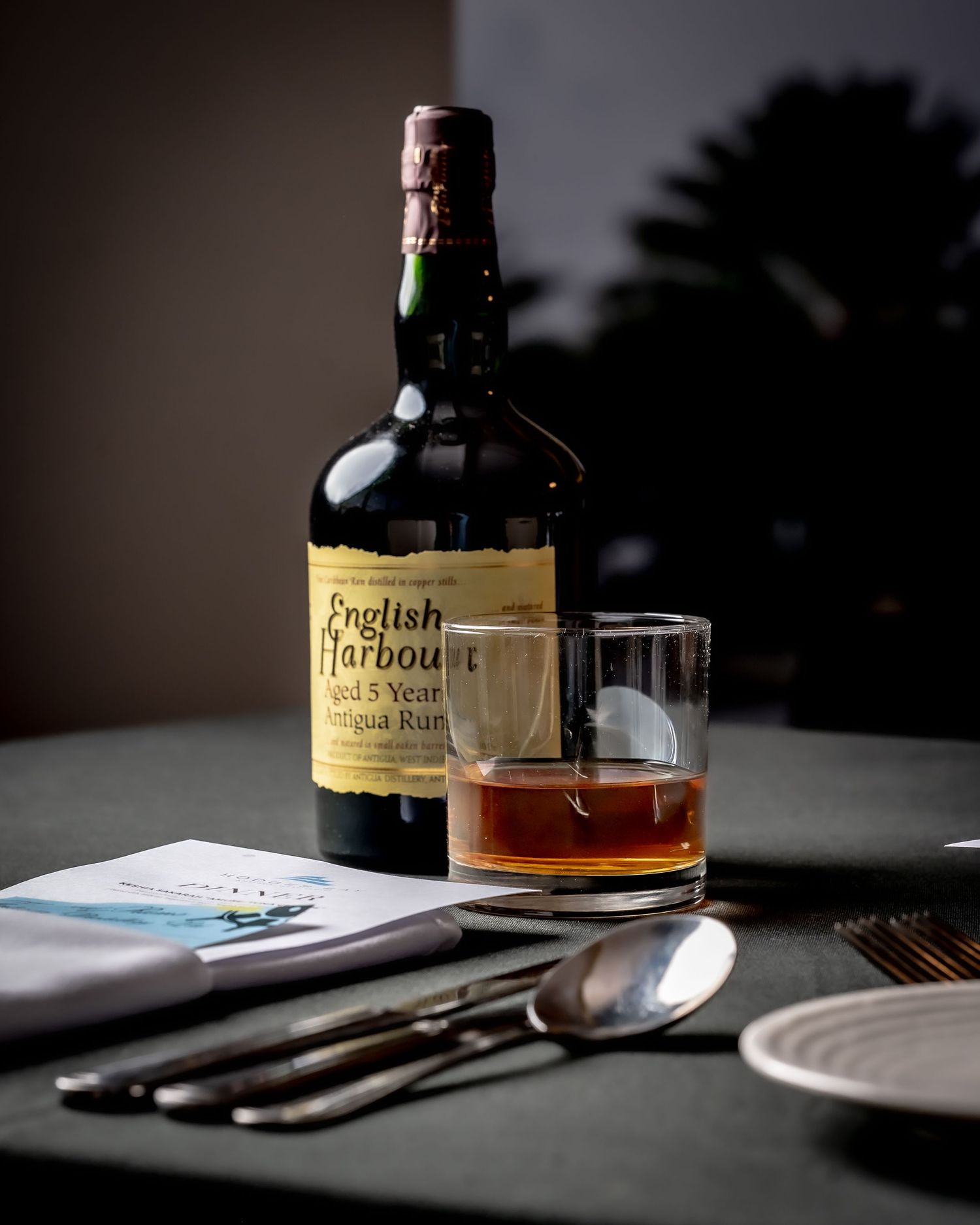 English Harbour Aged 5 Year Antigua Rum bottle beside a glass of rum on the rocks on a dark table.
