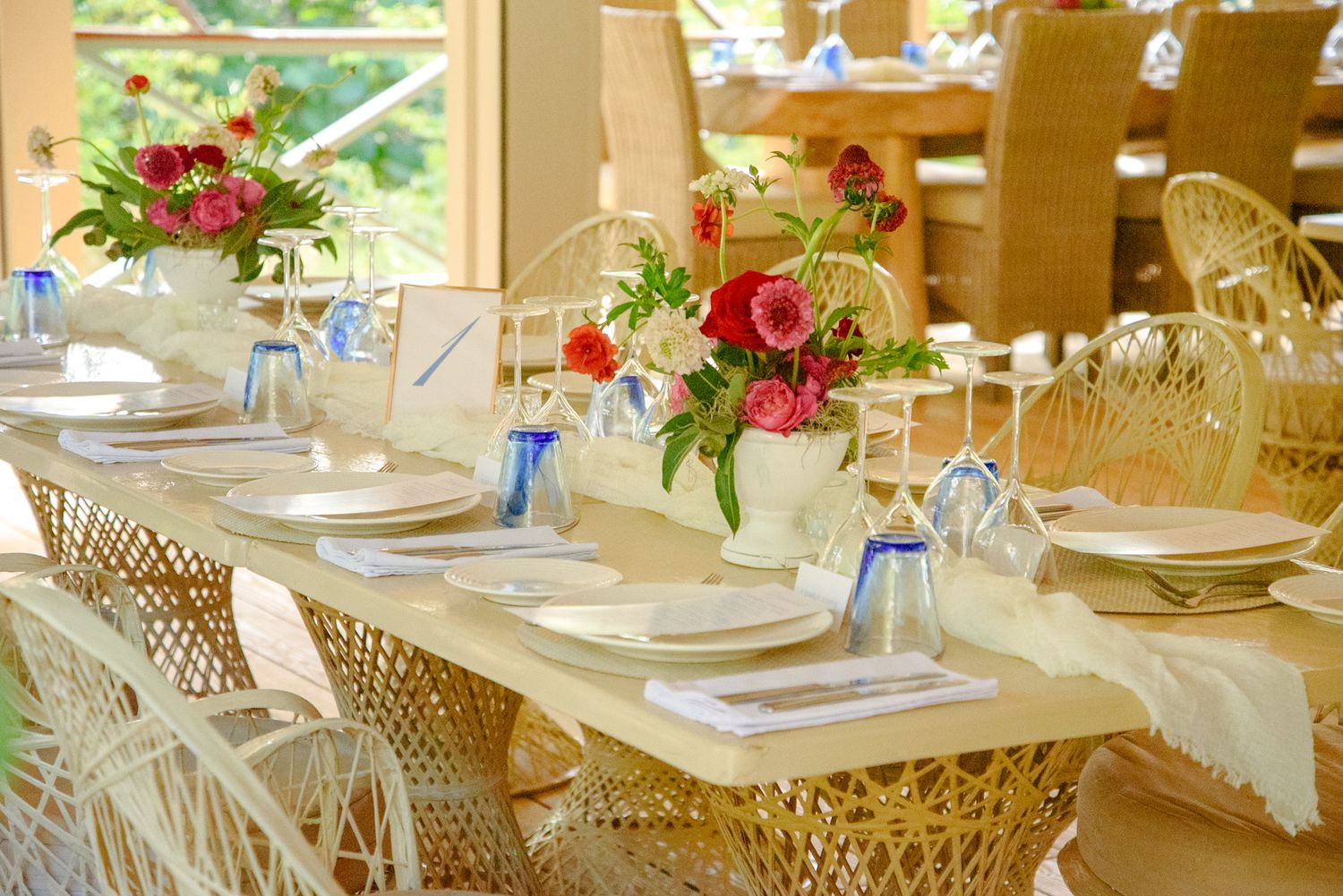 Elegant table settings with white linens, wicker chairs, and red floral centerpieces in a sunlit dining space.