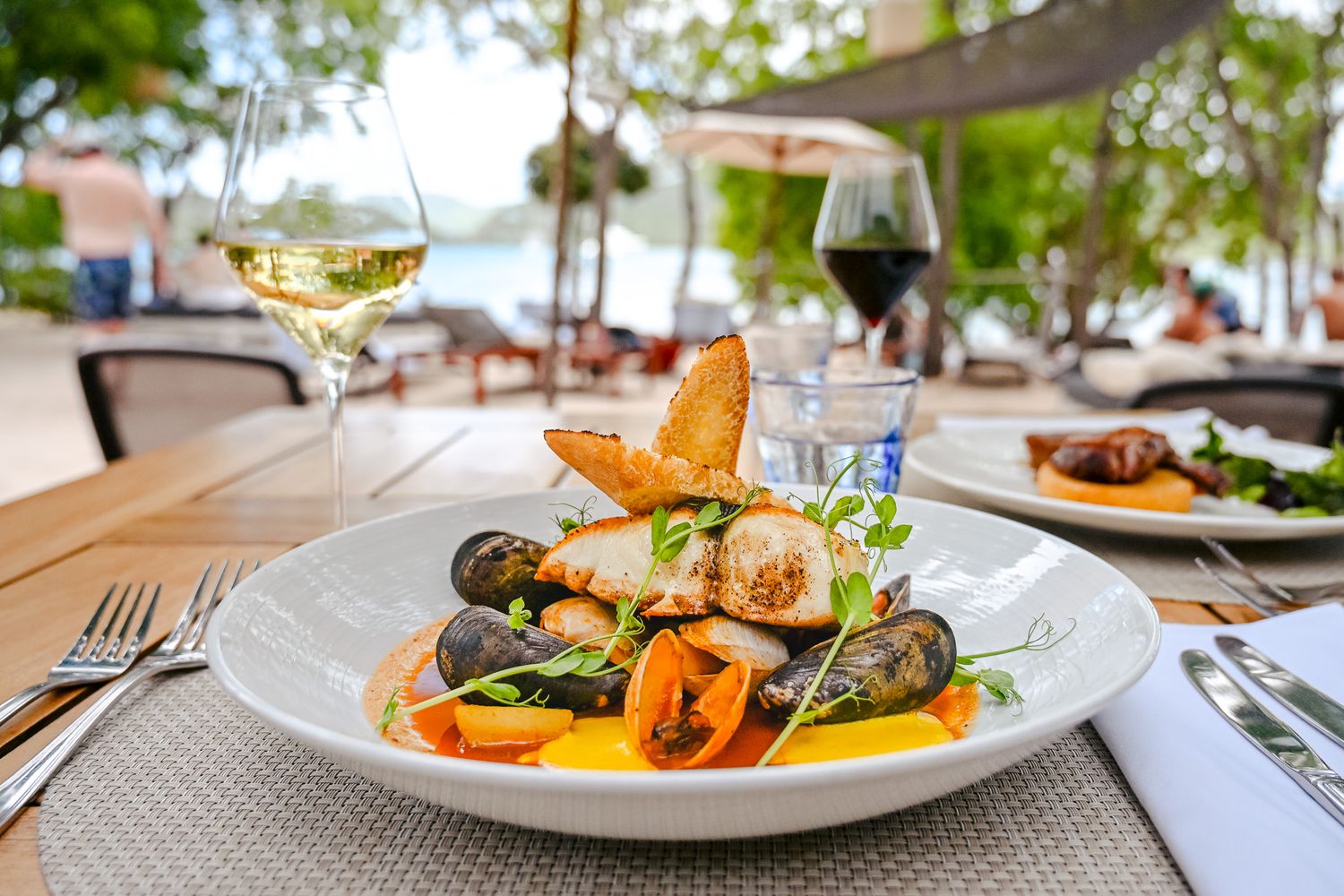 Gourmet seafood dishes with mussels and colorful vegetables arranged on white plates in an outdoor dining setting.