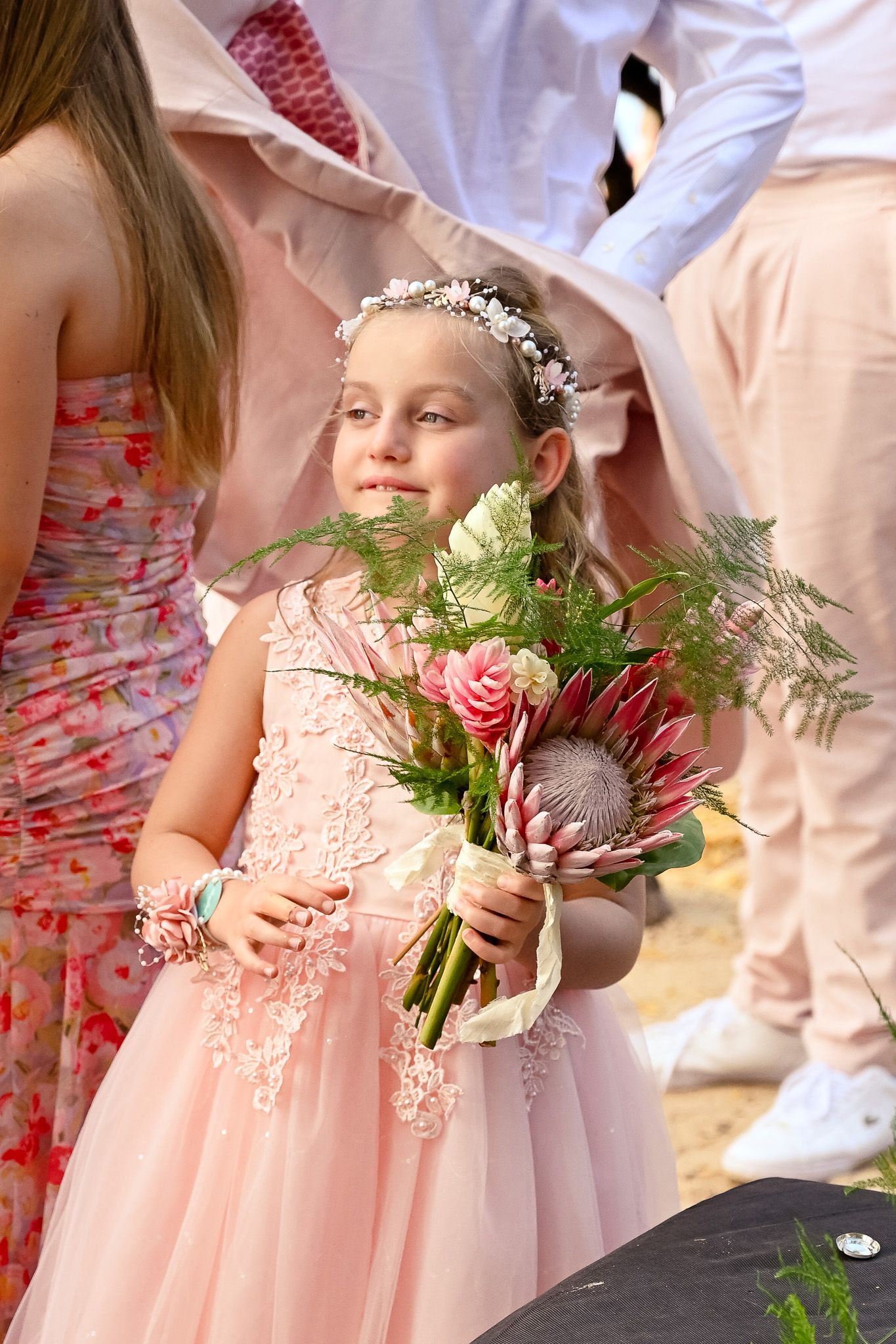 Young flower girl in a peach lace dress holds a pink floral bouquet with greenery at an outdoor wedding ceremony.
