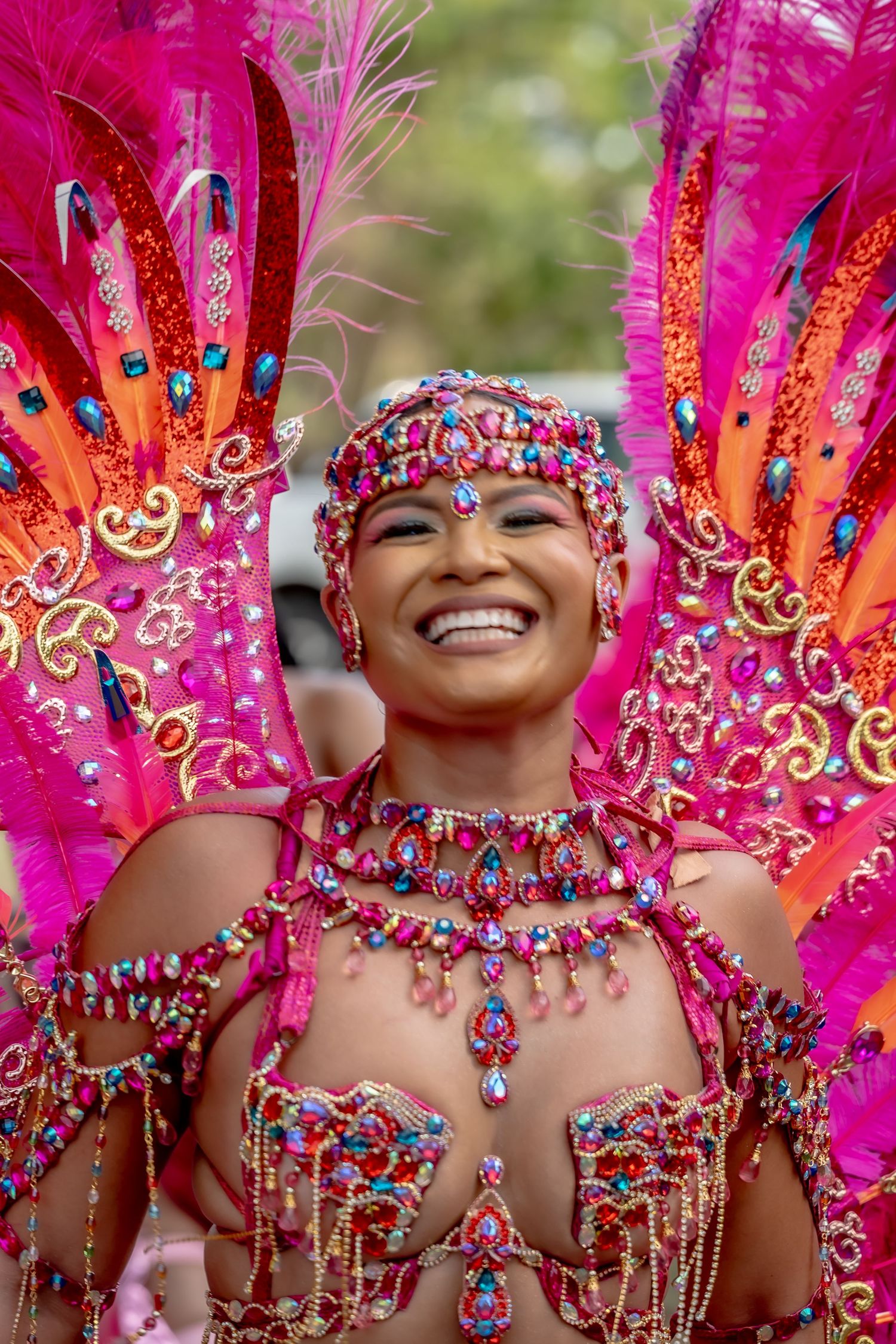 Smiling woman in vibrant pink and gold carnival costume with feathers and jewels at a festive parade.