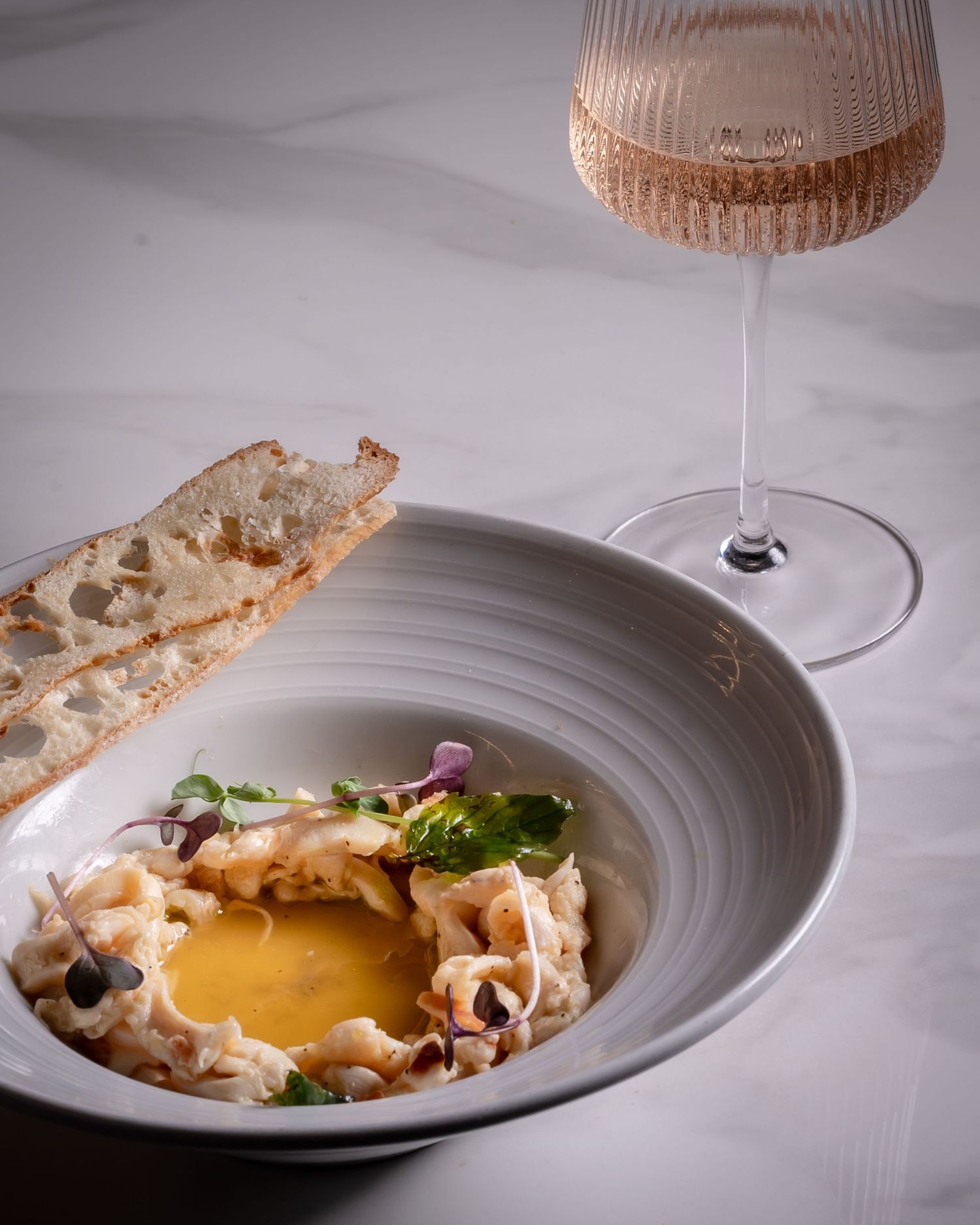 Elegant fine dining dish with creamy crab, golden egg yolk, microgreens, and toasted bread served in a gray bowl beside rosé wine.