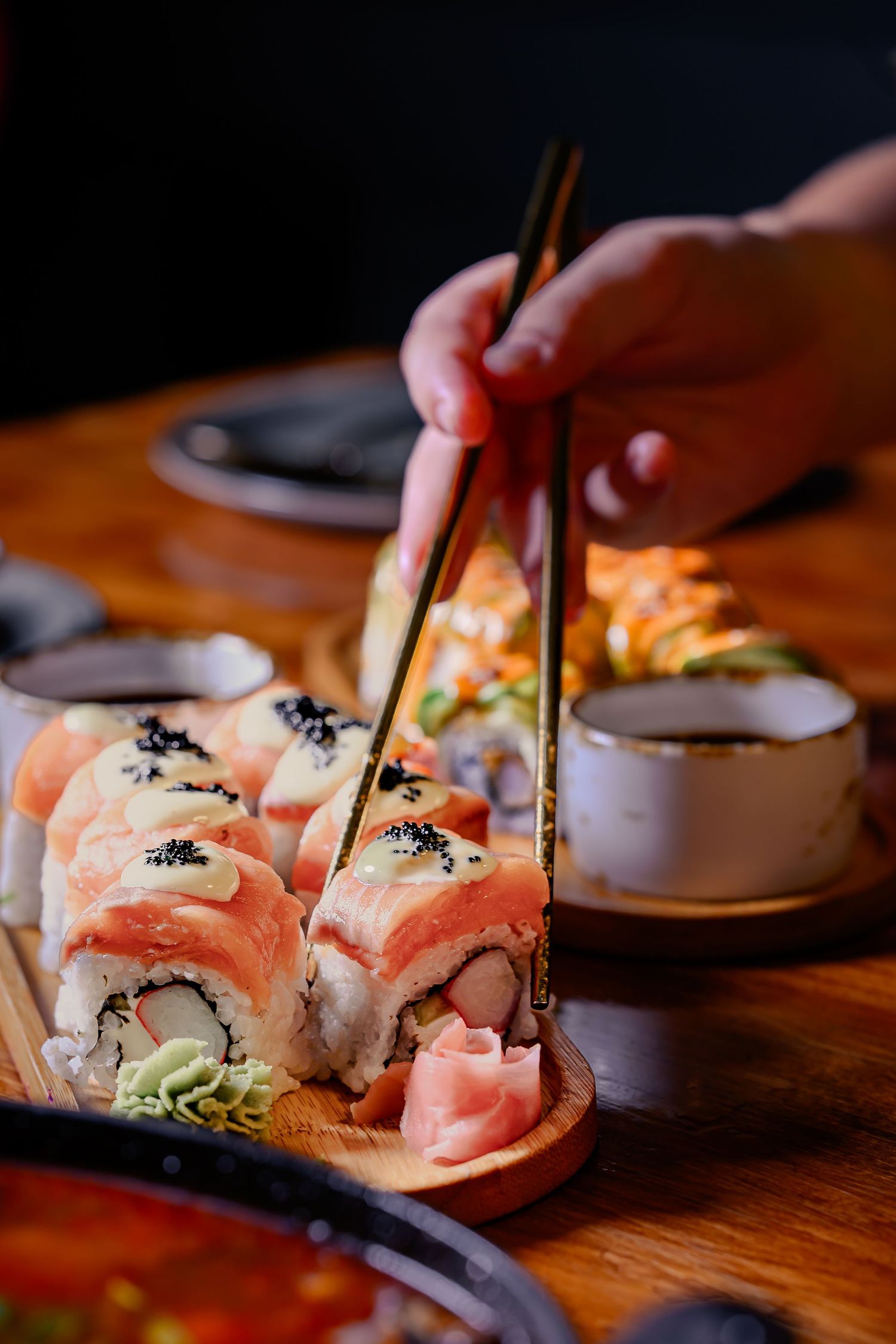 Hand picking salmon sushi roll with chopsticks from wooden platter, with soy sauce and pickled ginger.