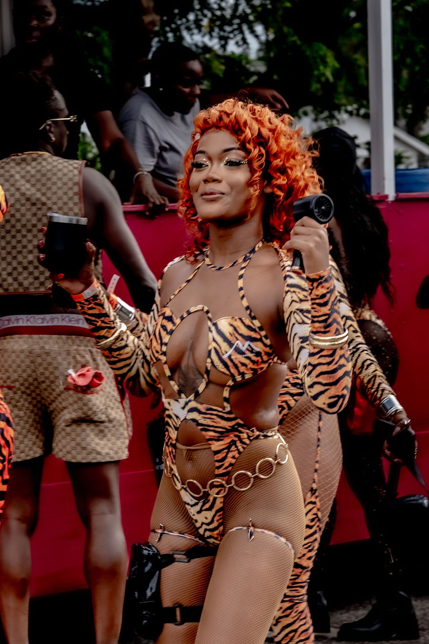 Woman in tiger-print carnival costume with orange curly hair holds microphone at vibrant outdoor festival.