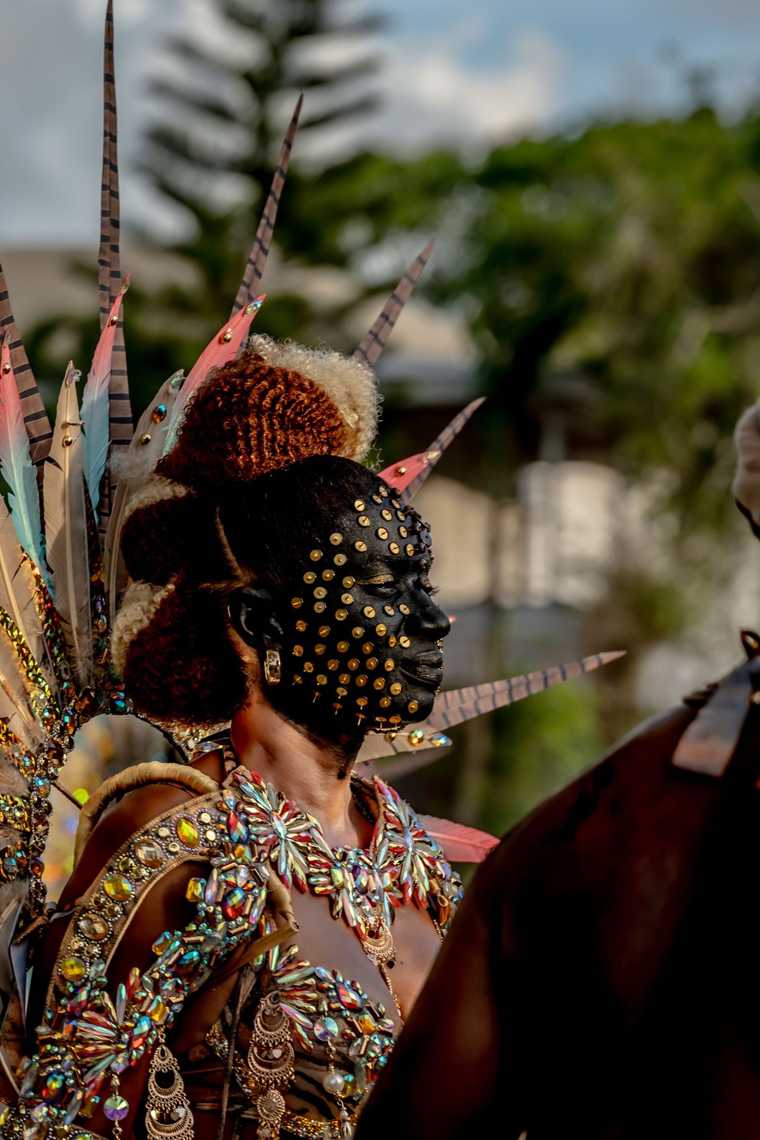 Carnival performer with black face paint, gold dots, feathered headdress, and jeweled costume at a colorful festival parade.