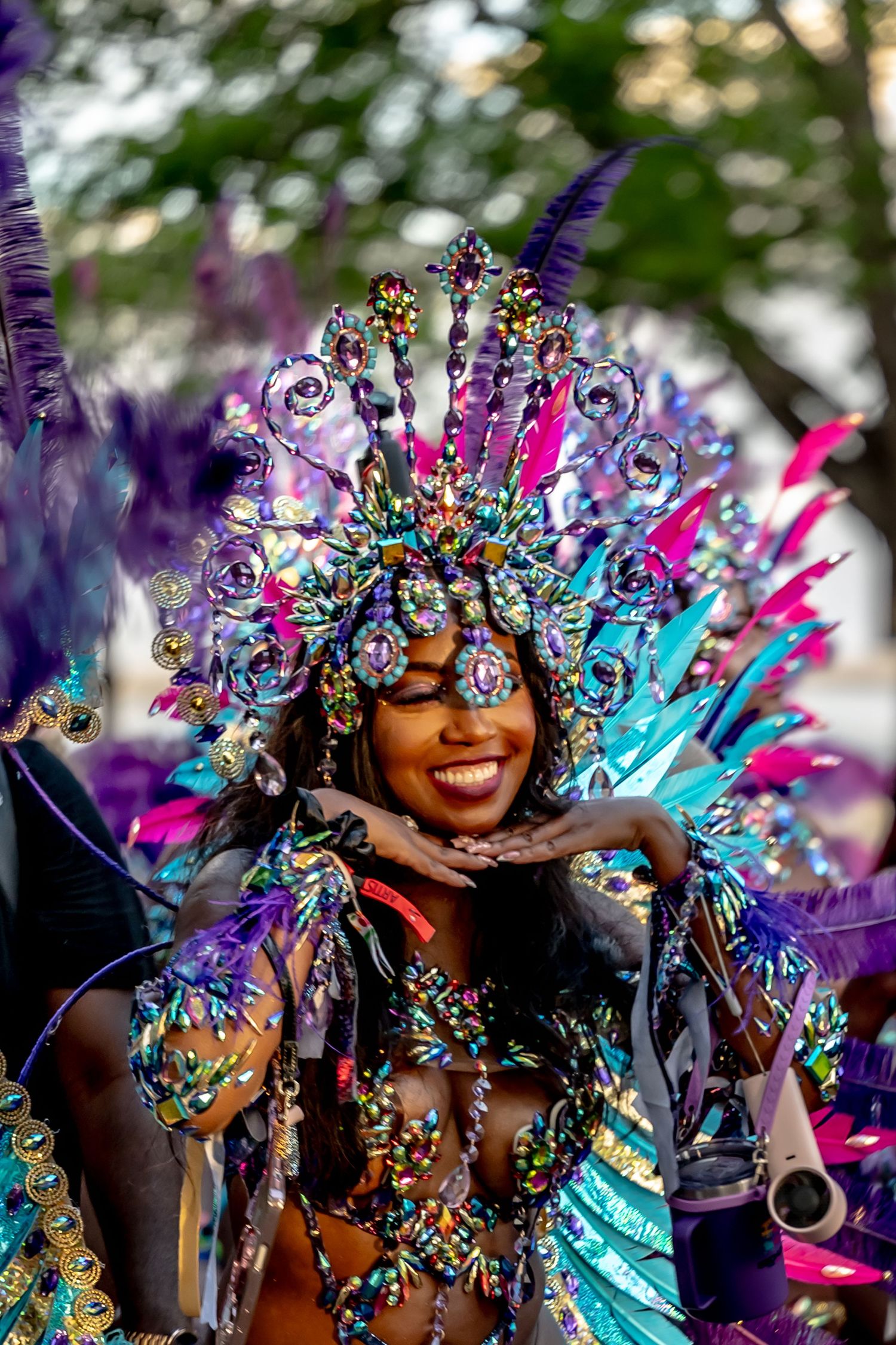 Smiling woman in vibrant turquoise and purple carnival costume with ornate feathered headdress blowing a kiss.