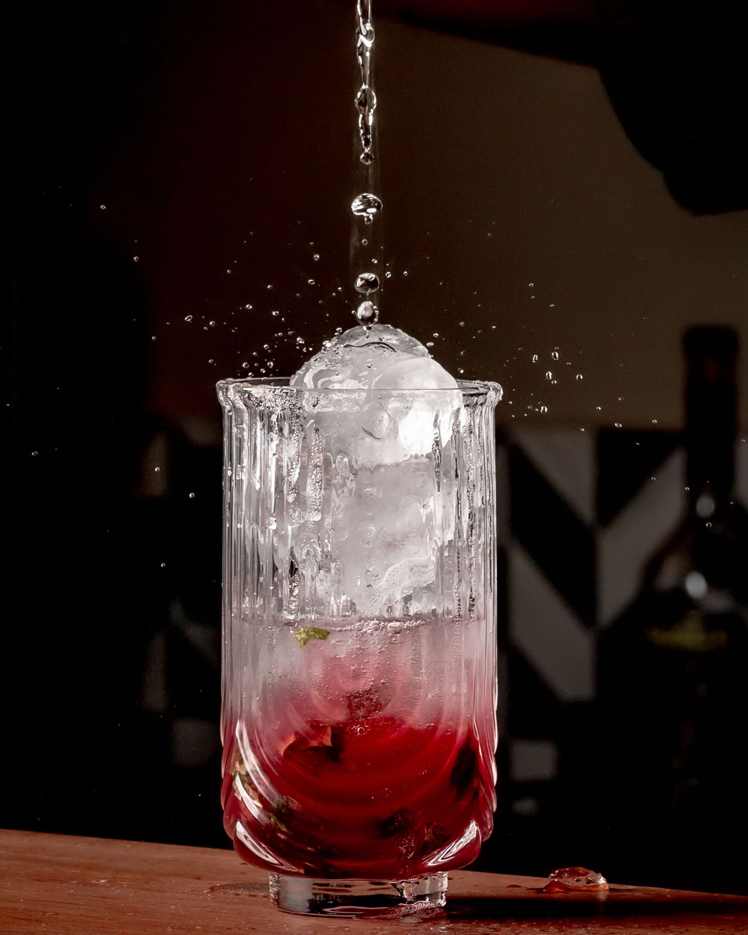 Water pouring over a large ice sphere in a ribbed crystal glass with a vibrant red cocktail base on a wooden bar.
