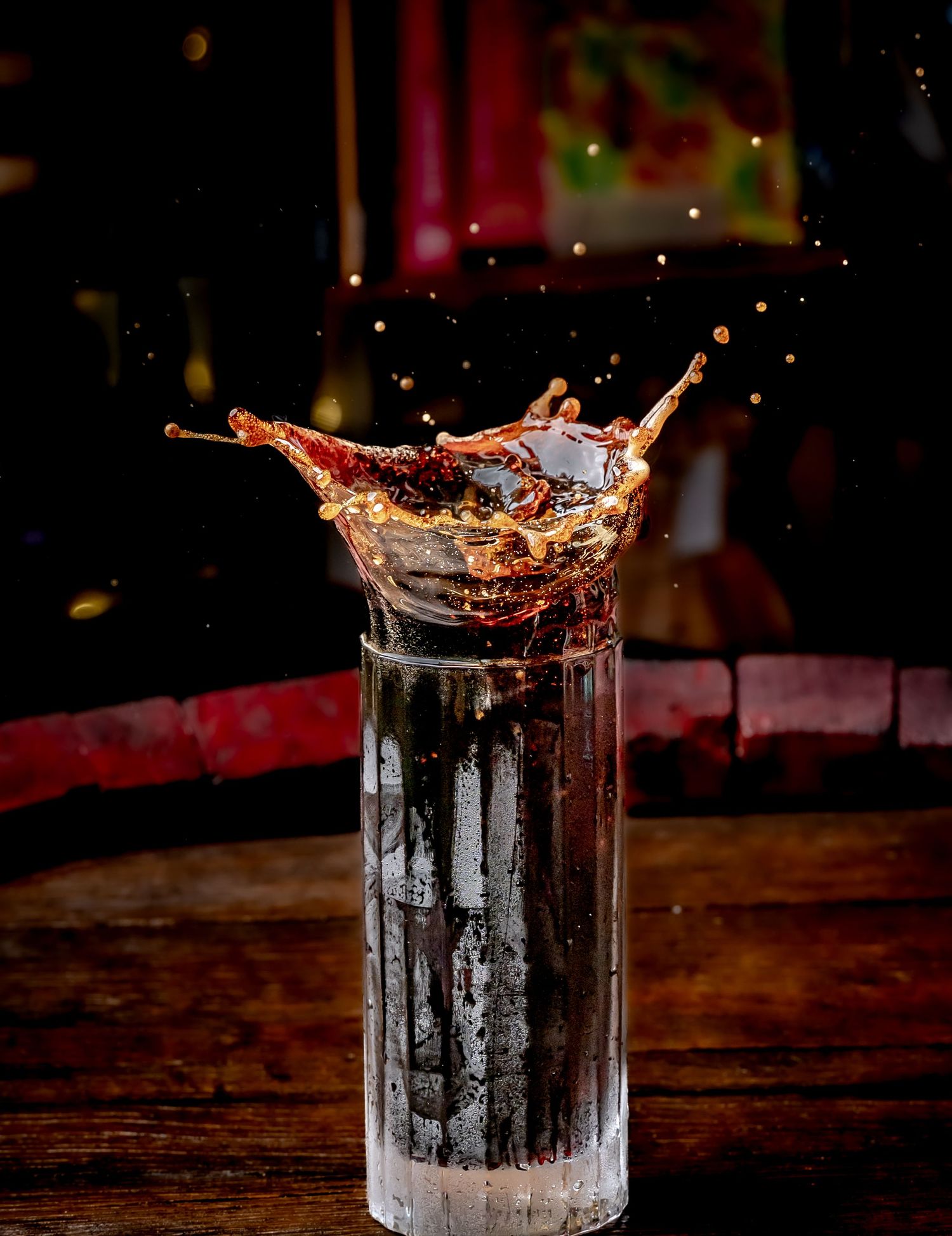 A dramatic liquid splash erupts from a tall glass of dark cola on a rustic wooden table in moody bar lighting.
