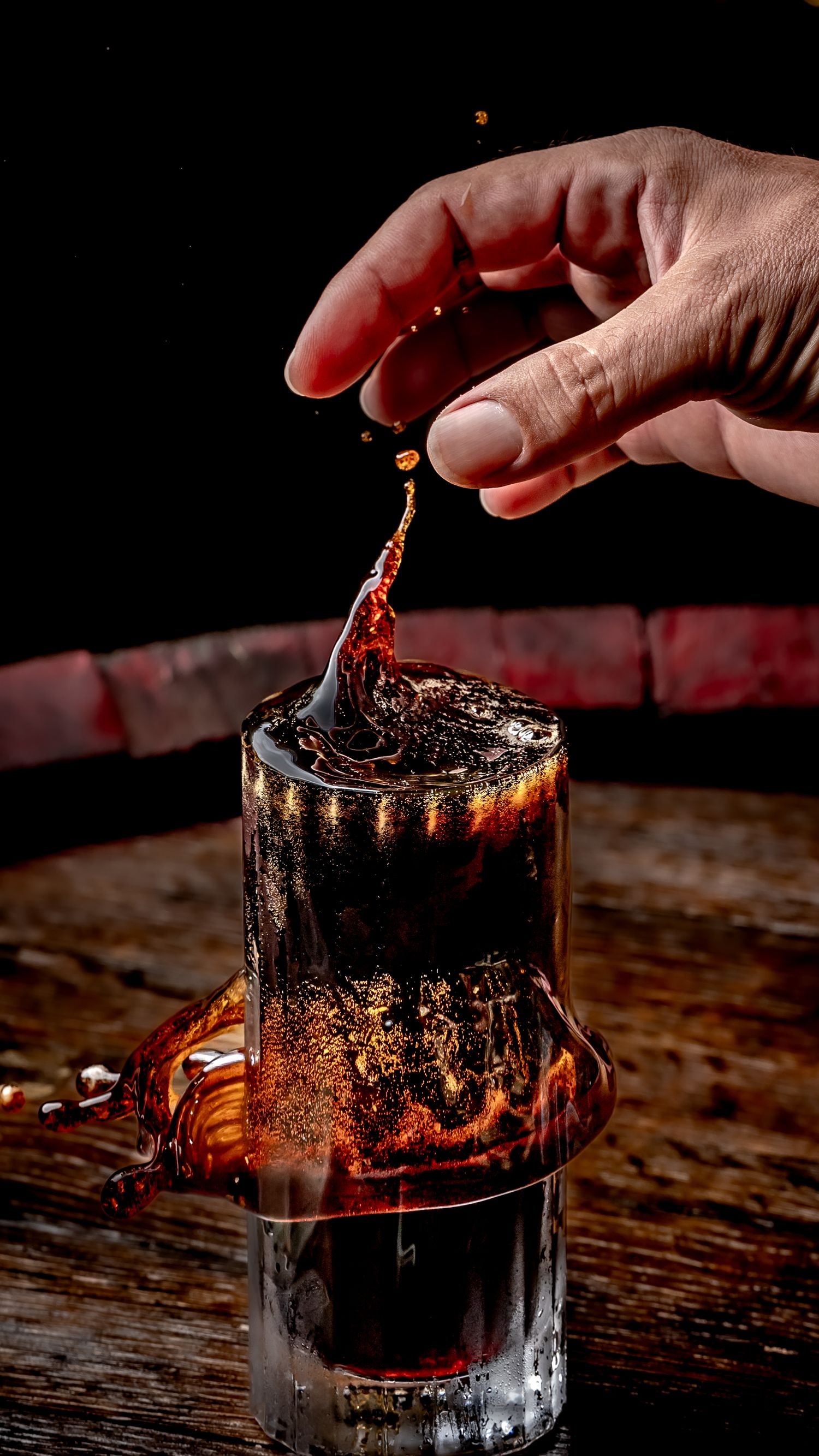 Hand dropping liquid into a dark whiskey cocktail glass on a wooden barrel, creating a dramatic splash effect.