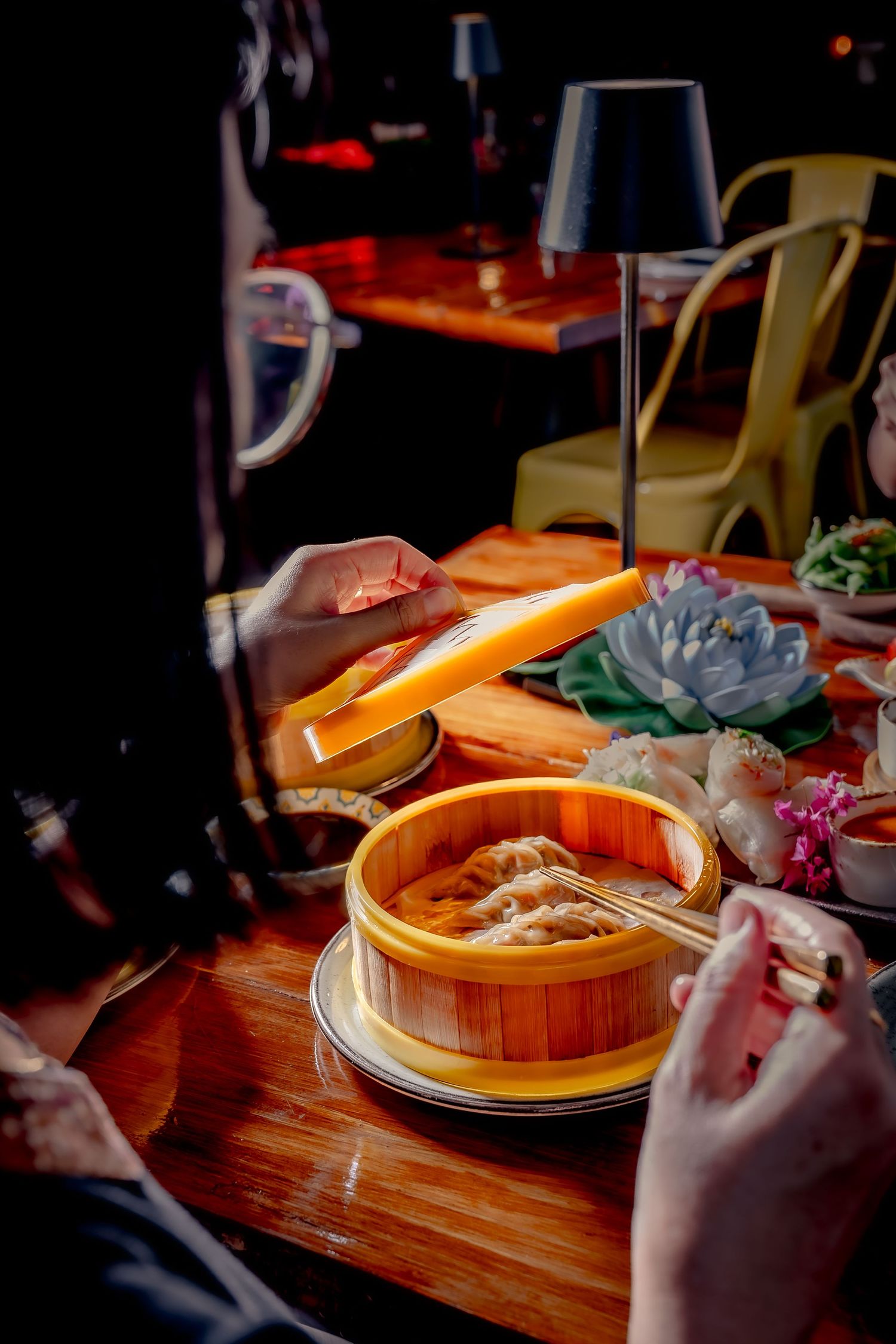 Hands serving dim sum dumplings from a bamboo steamer basket on a wooden table in a dimly lit restaurant.