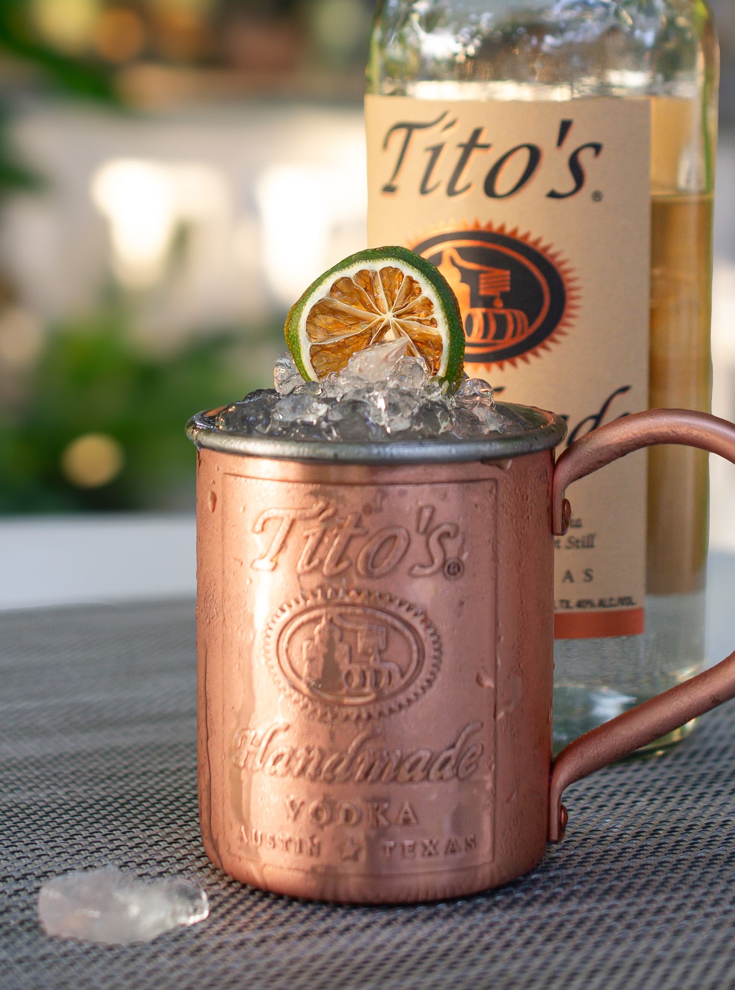 A series of copper Moscow Mule mugs filled with ice and garnish sit on a dark surface with Tito's vodka bottle visible.