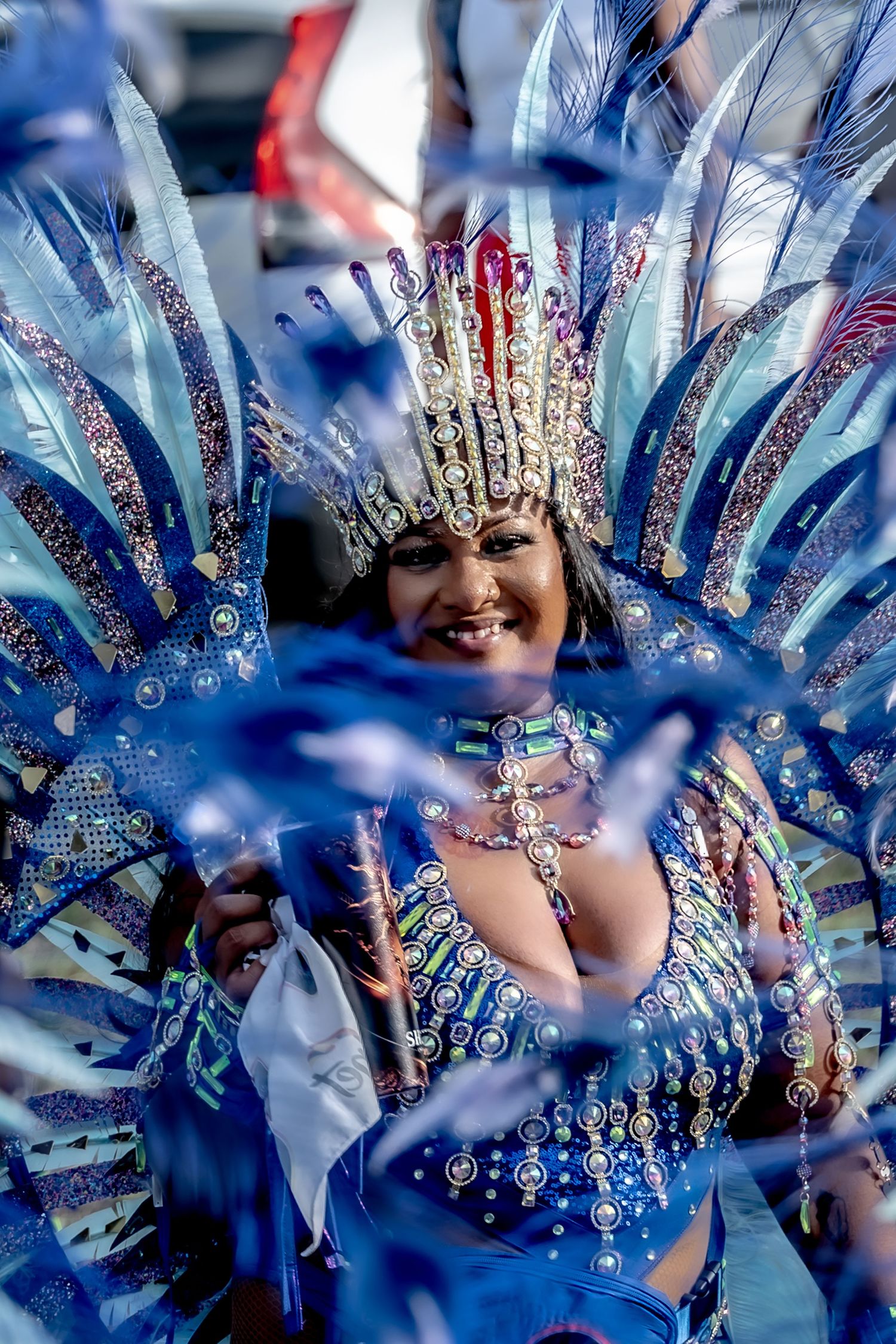 Smiling woman in elaborate blue feathered carnival costume with jeweled headdress at a festive parade.