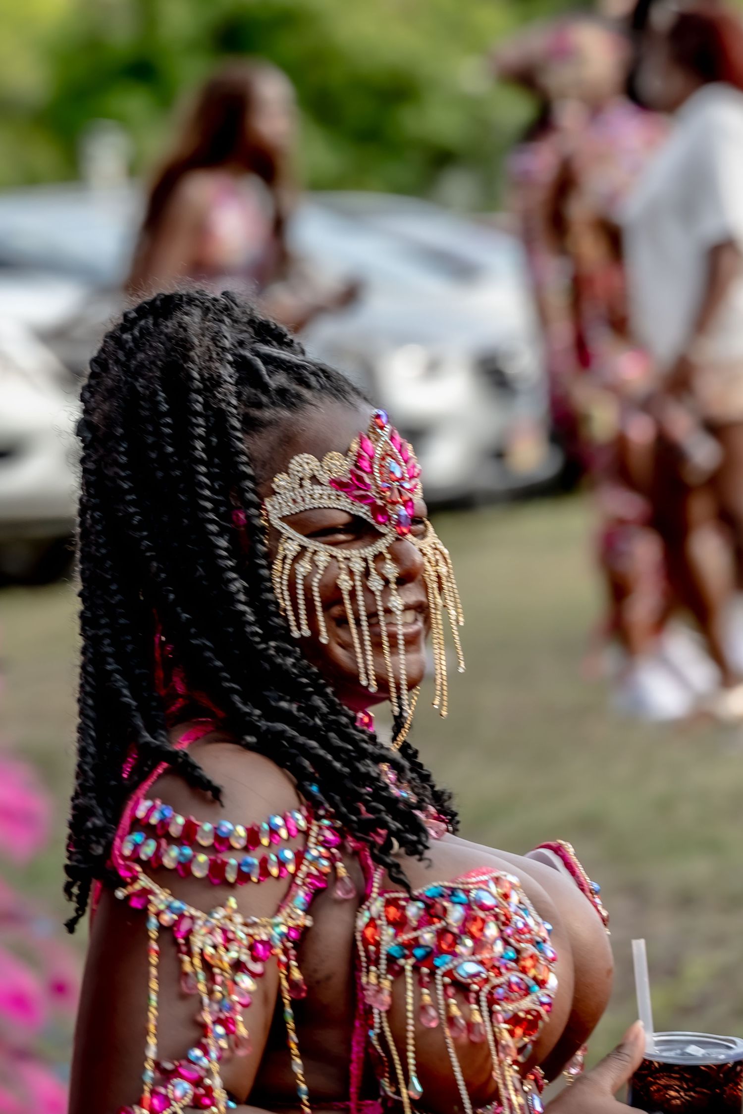 A woman in vibrant pink carnival costume with jeweled mask and braided hair at a festive outdoor celebration.