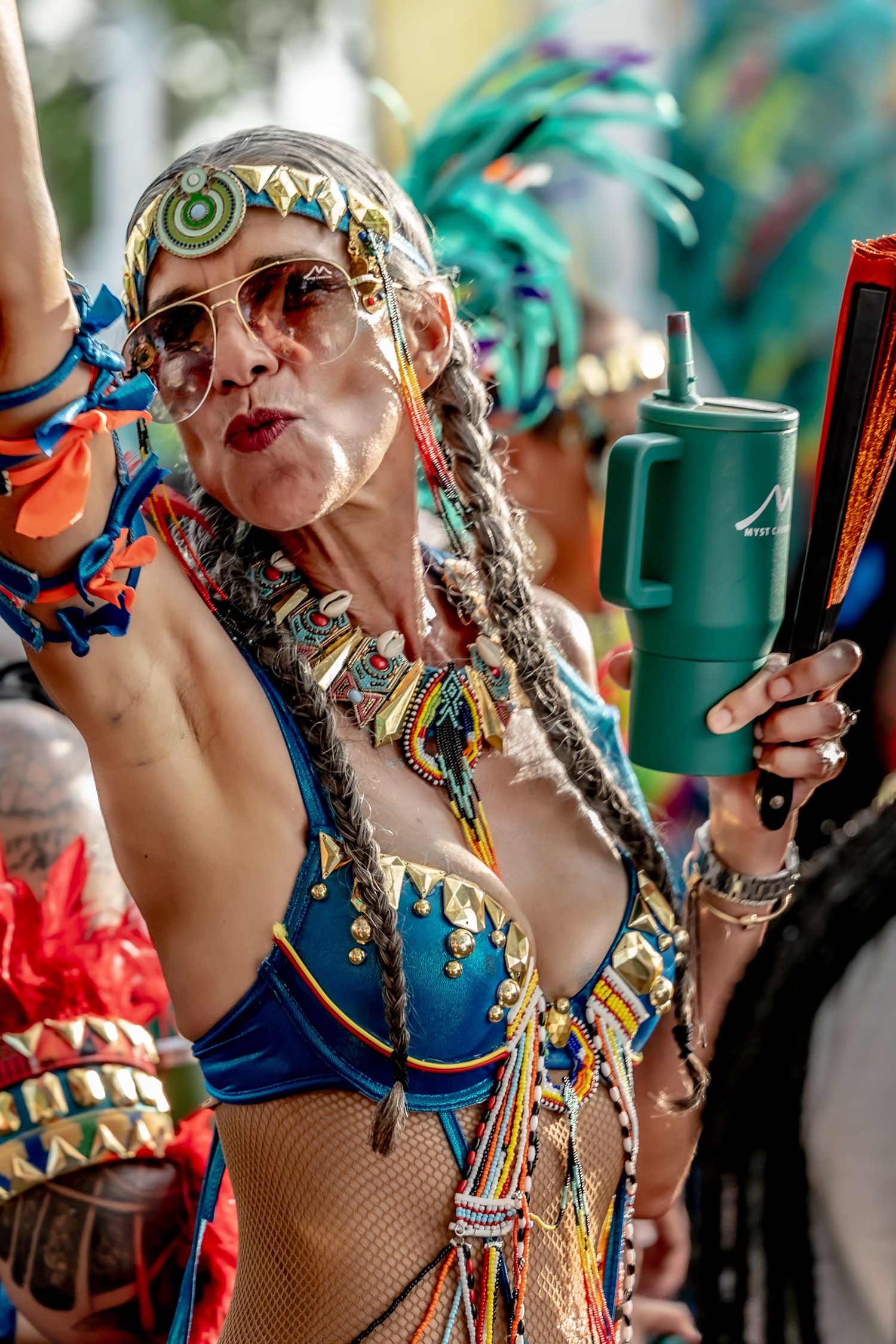 Woman in colorful carnival costume with braids, beaded headpiece, sunglasses, holding a tumbler at a festive parade.
