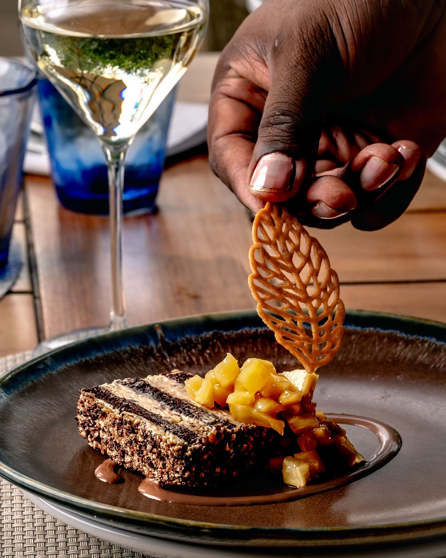 Hand placing a decorative leaf tuile on a layered chocolate cake with mango compote on a dark plate beside white wine.