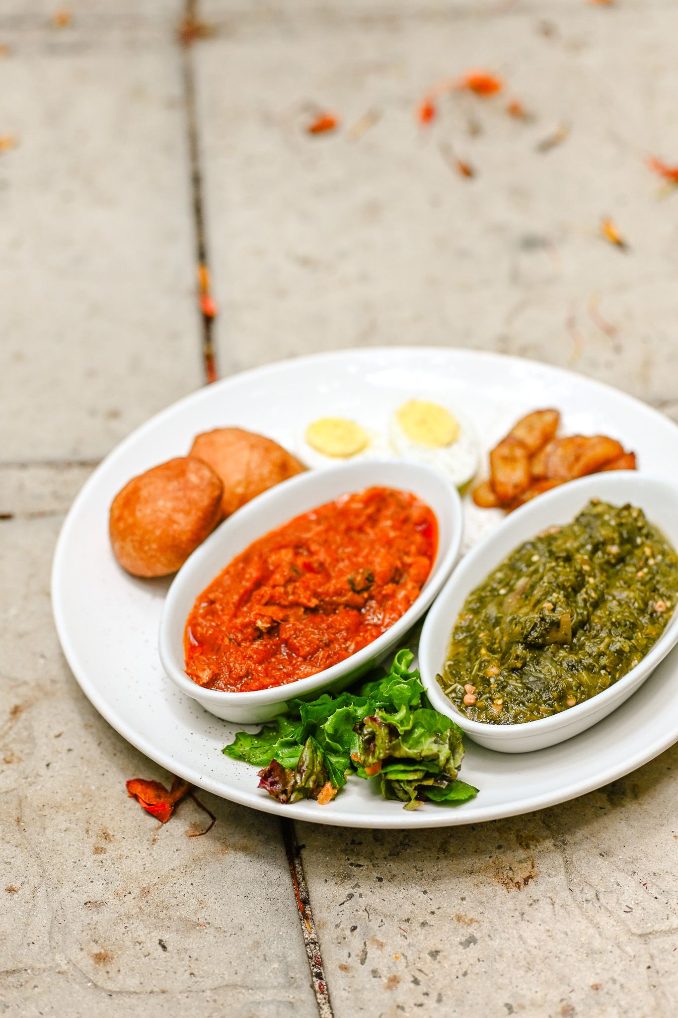 White plate with red and green chutney dips, fried bread, boiled egg slices, and fresh herbs on rustic surface.