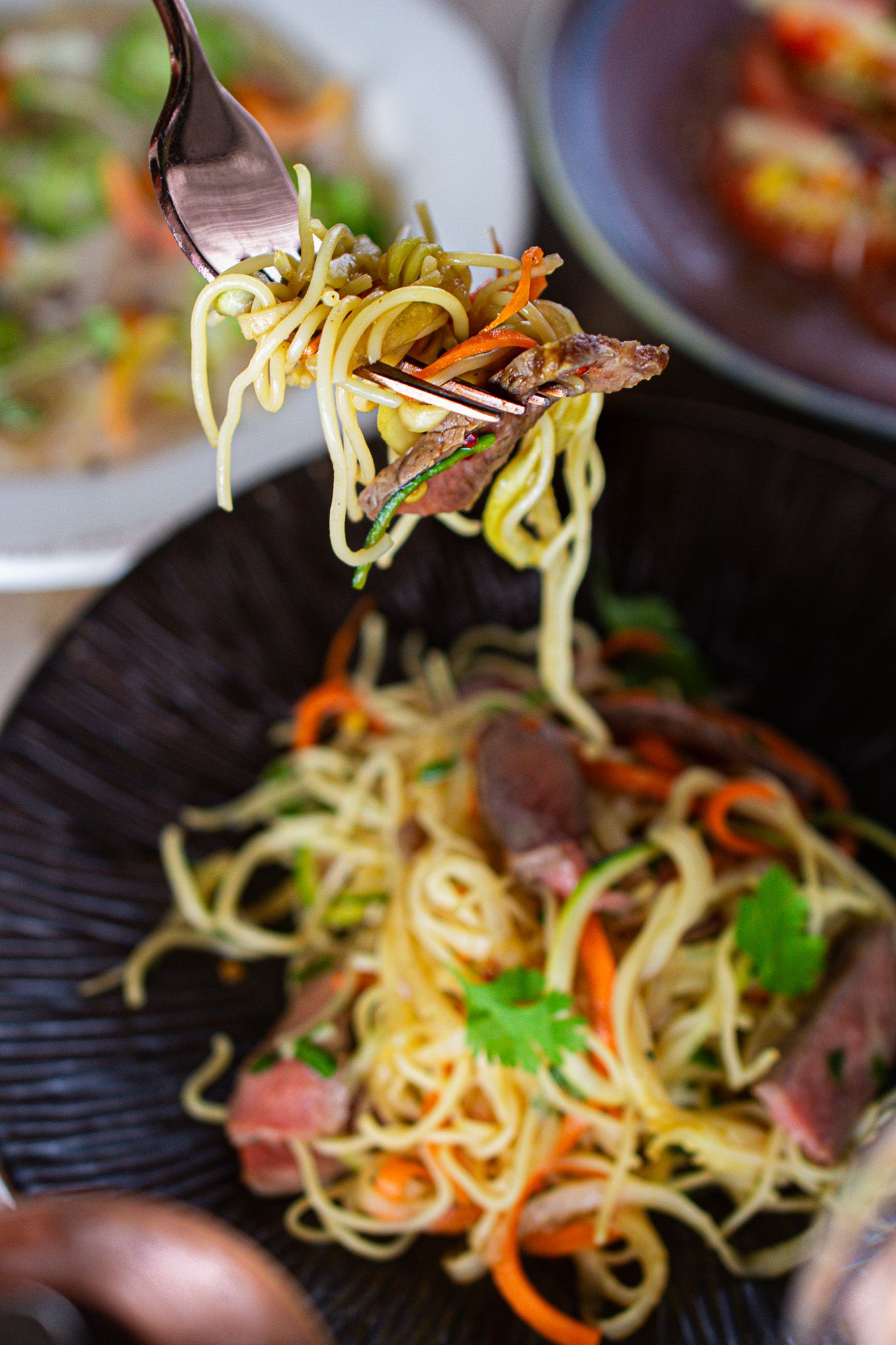 A fork twirls Asian noodles with sliced beef, carrots and herbs from a black plate in a close-up shot.