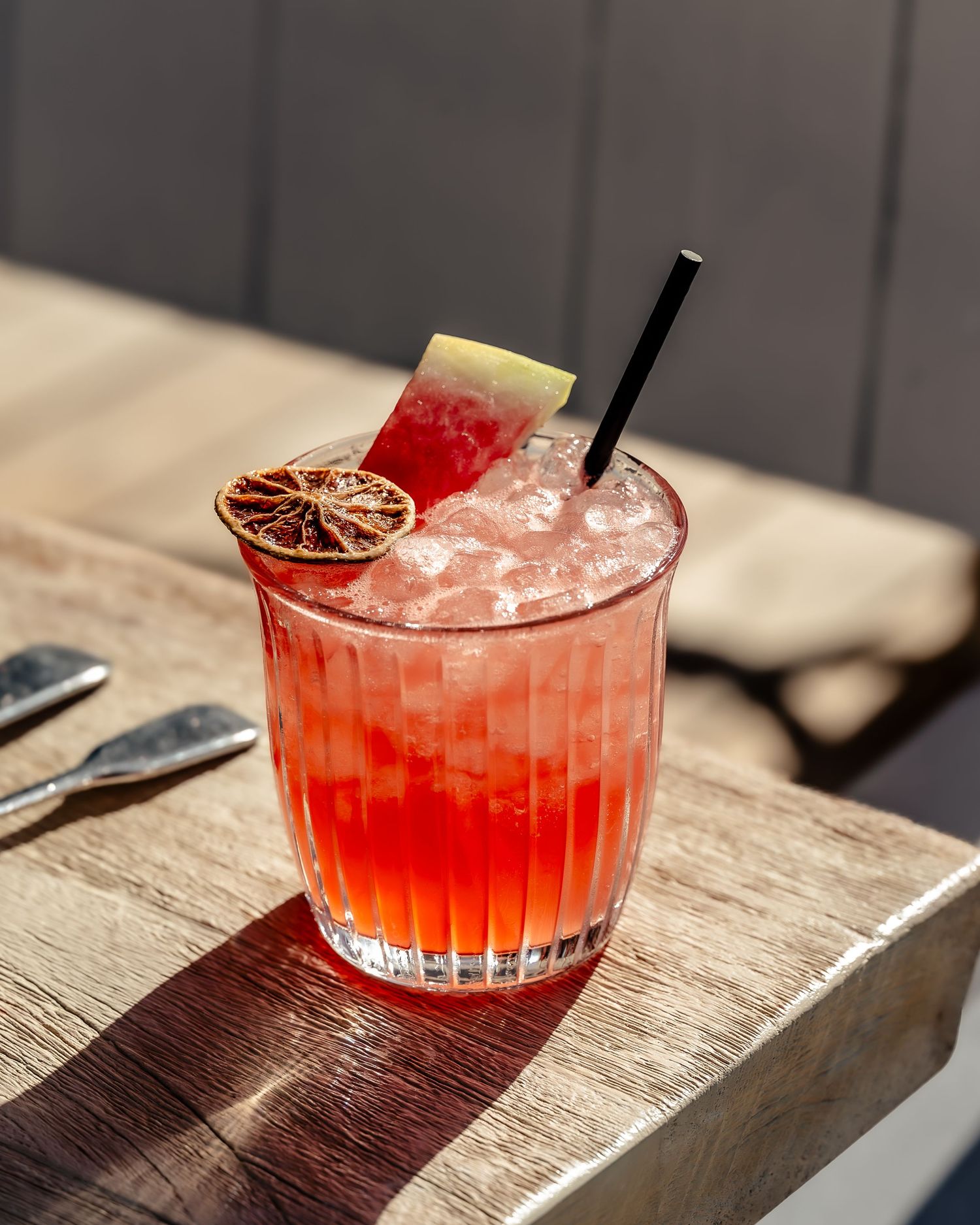 A vibrant red cocktail on crushed ice garnished with dried citrus and watermelon slice in a ribbed glass outdoors.