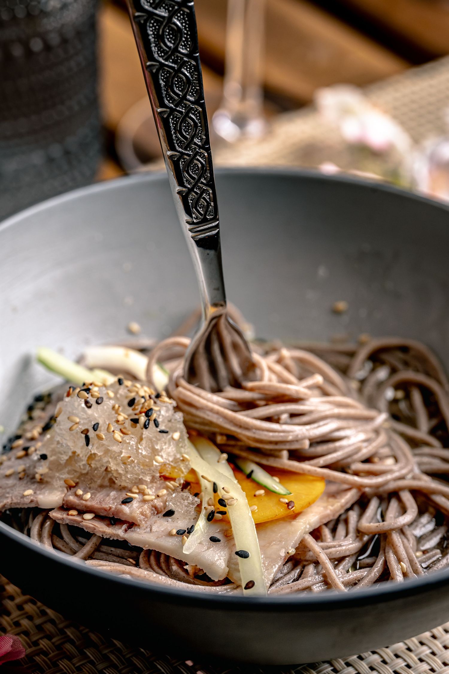 A gray bowl of soba noodles topped with sesame seeds, vegetables, and a decorative fork.