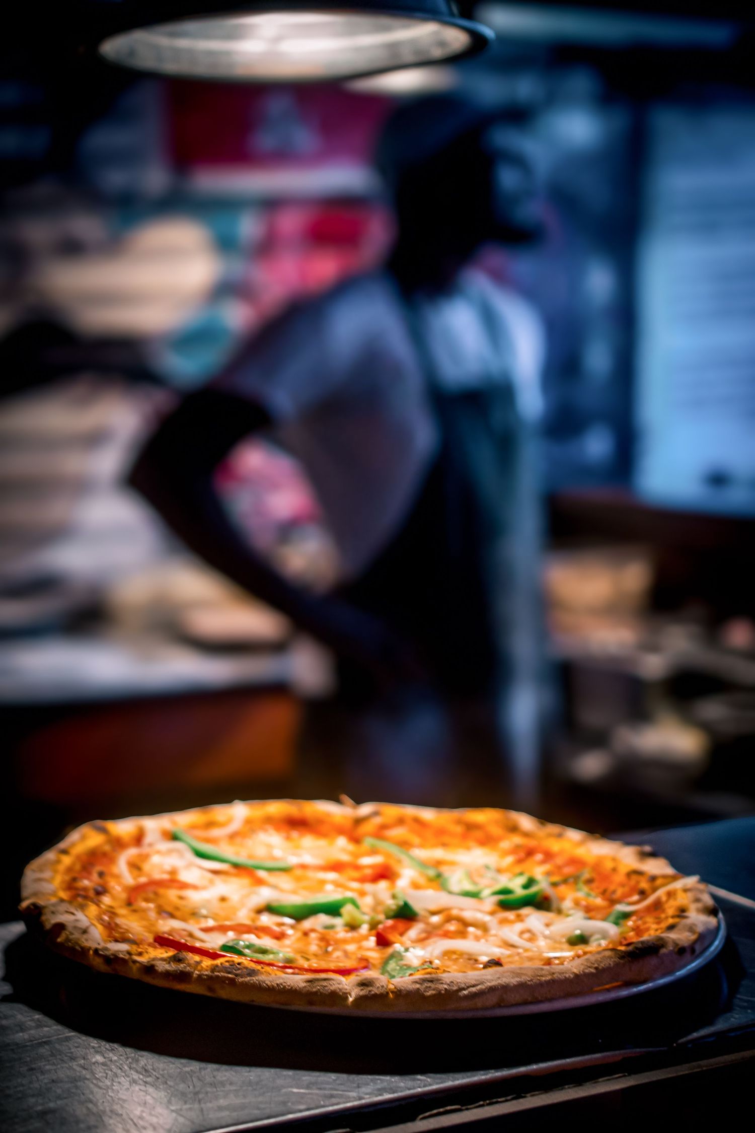 Freshly baked pizza with melted cheese and green peppers on a wooden board in a dimly lit restaurant kitchen.