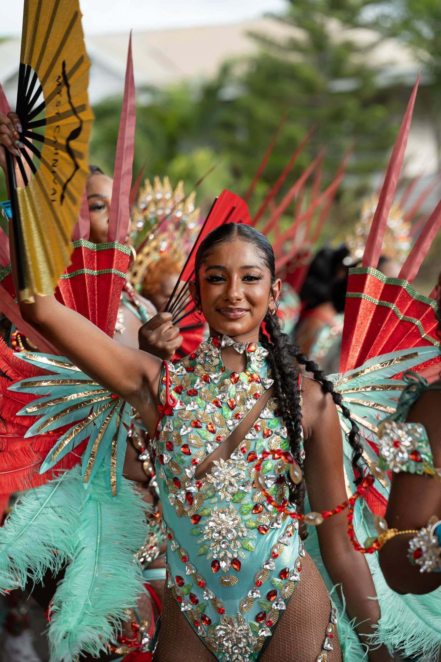 Smiling woman in vibrant green and red jeweled carnival costume with feathers and fans at a colorful parade.