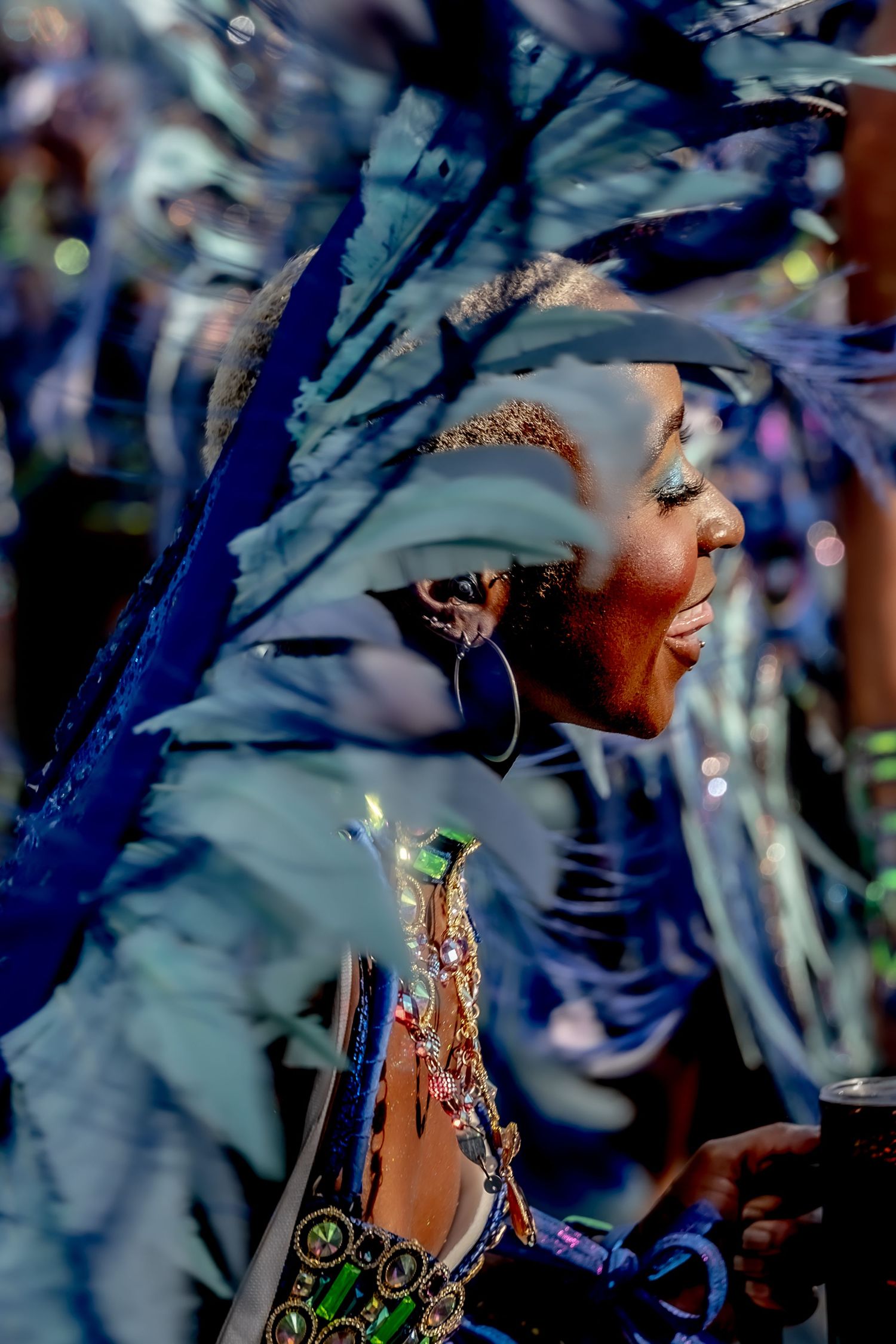Woman in vibrant blue feathered carnival costume with hoop earrings and jeweled accessories at a festival parade.