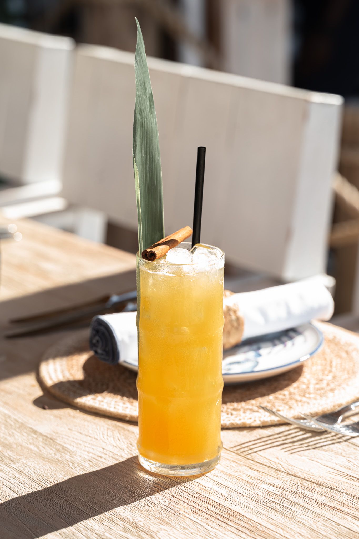 A tropical cocktail with orange juice and foam garnished with a palm leaf sits on a wooden table in bright sunlight.