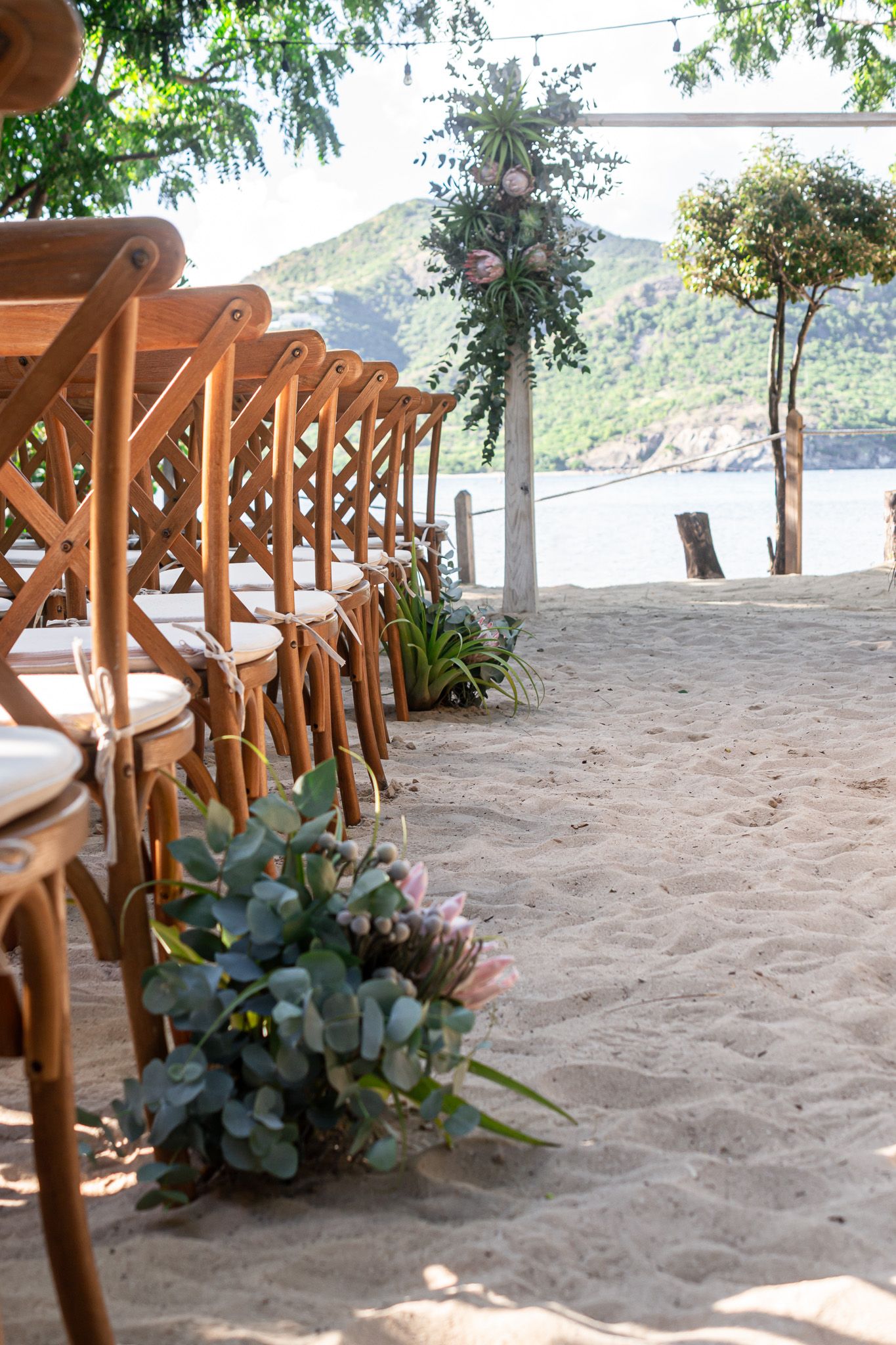 Wooden cross-back chairs arranged on a sandy beach with tropical plants and ocean views in the background.