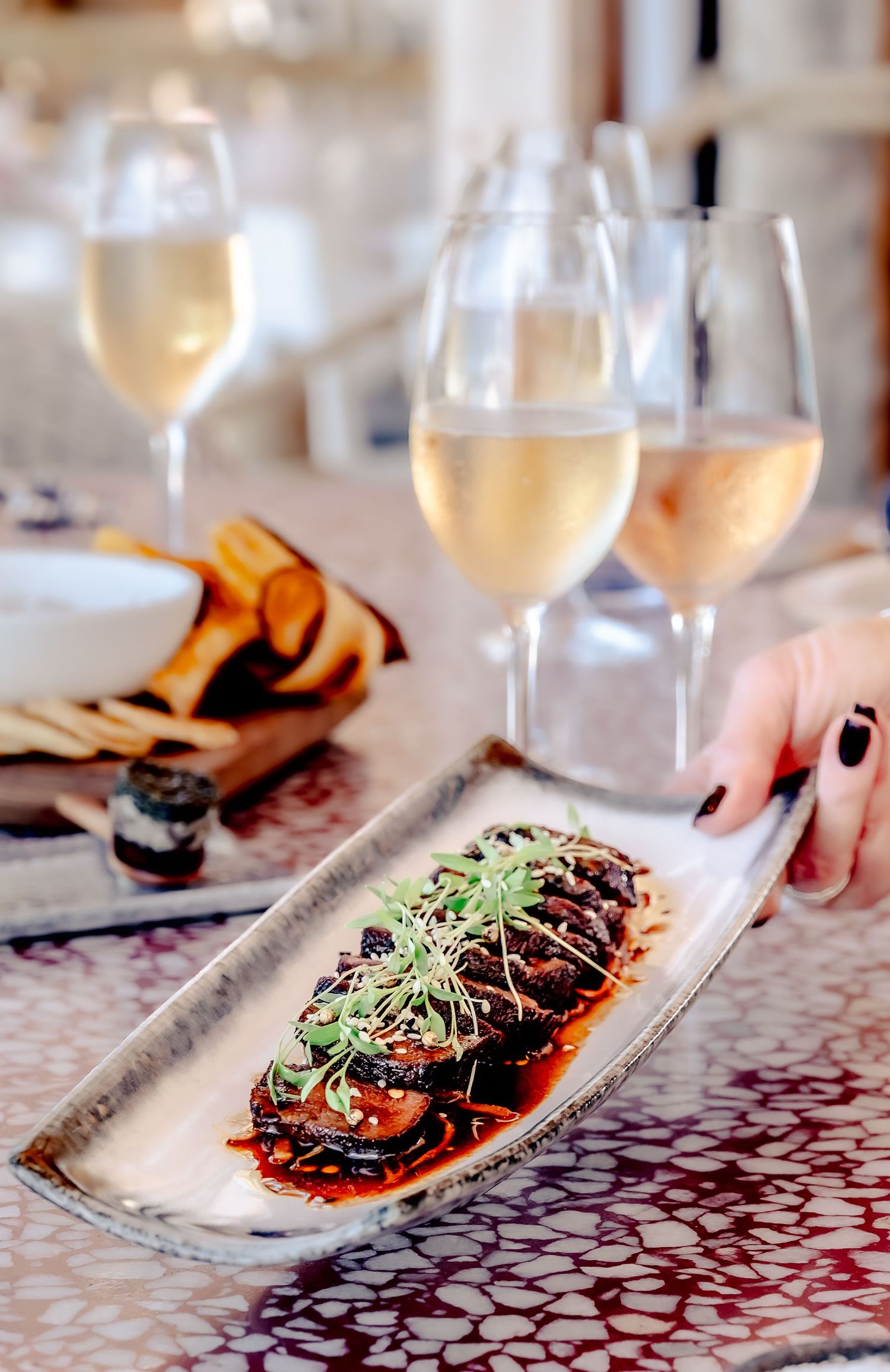 Elegant restaurant dish of braised eggplant with microgreens and sauce, served alongside two glasses of white wine.