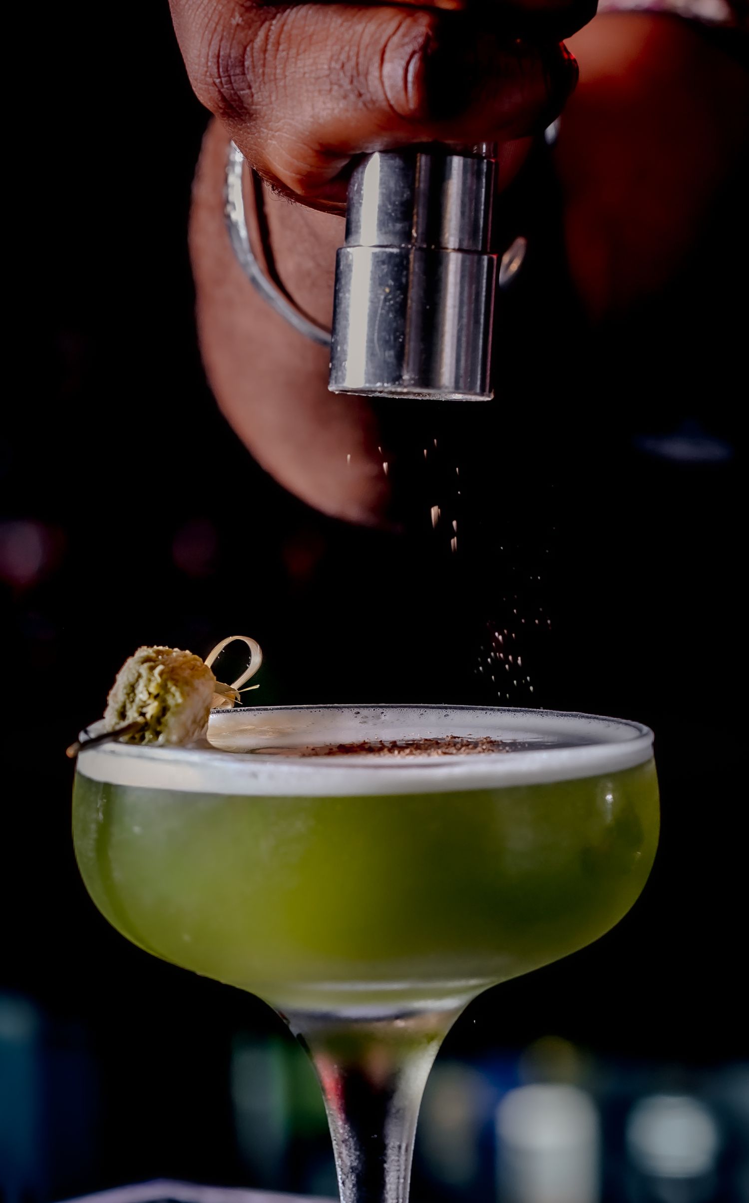 Bartender dusting spice over a green cocktail in a coupe glass with a garnish, dark moody bar setting.