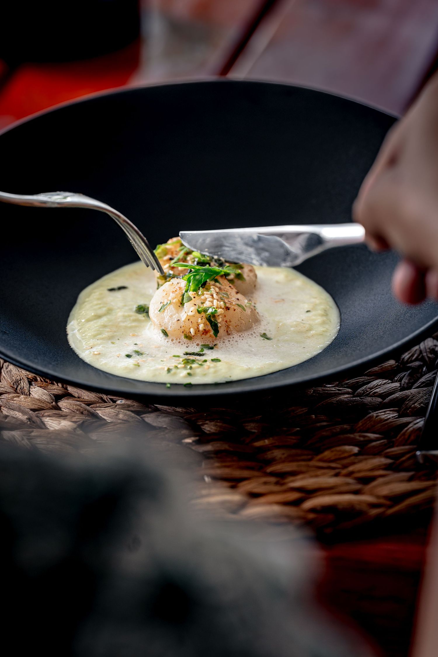 Elegant seared scallop in creamy sauce, garnished with herbs, served in a black bowl with silverware.