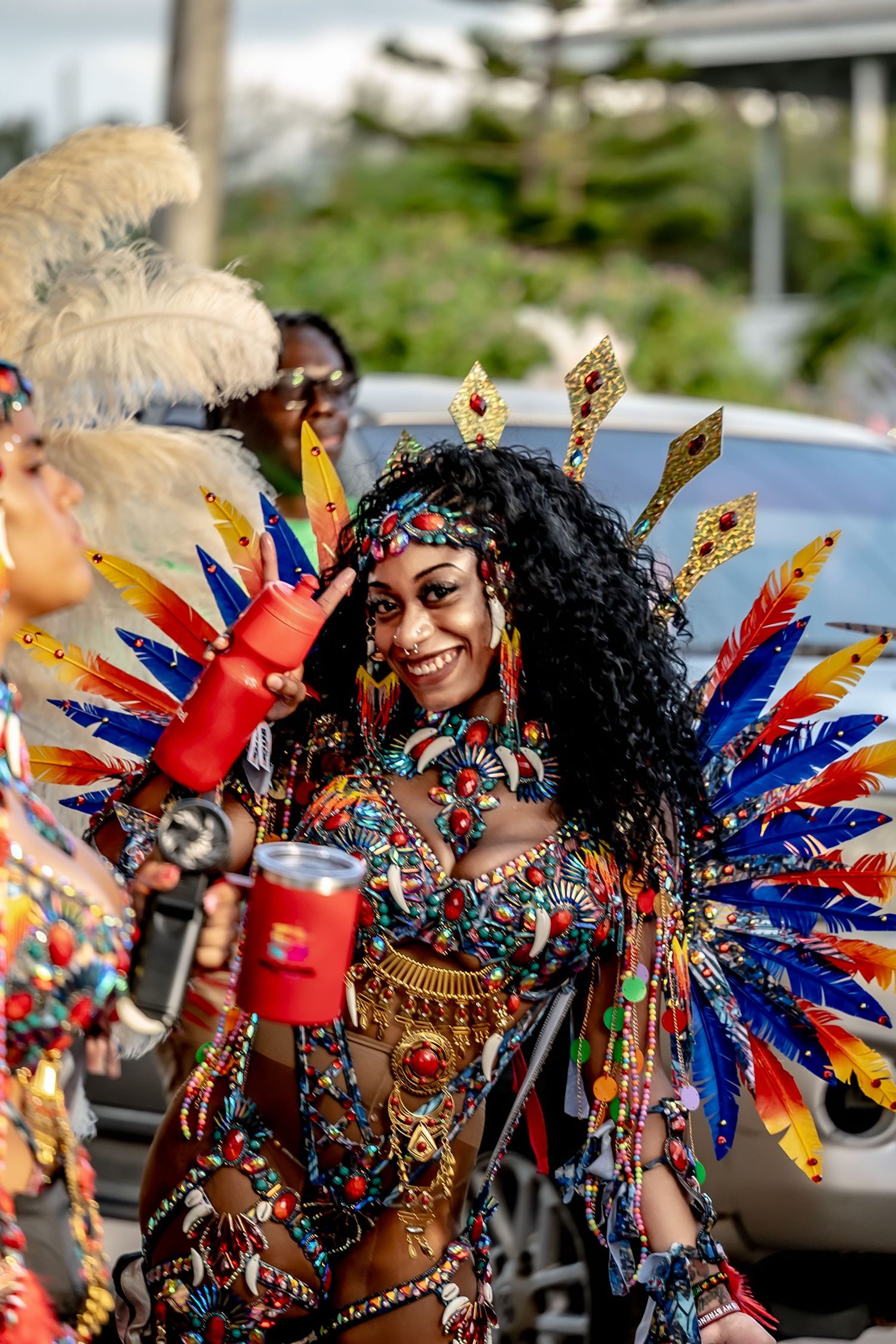 A smiling woman in a vibrant carnival costume with colorful feathers and jeweled accessories at a festive parade.