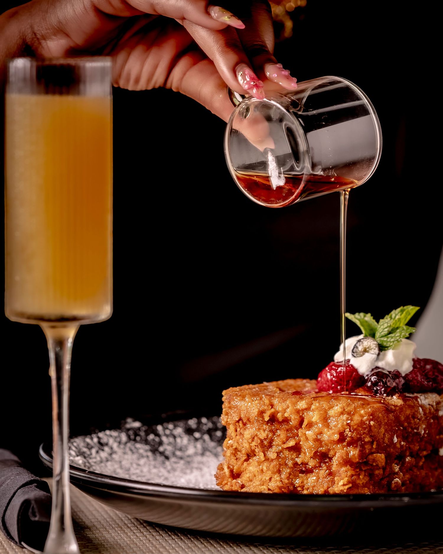 Hand pouring syrup over a golden French toast topped with berries and cream, served with a mimosa.