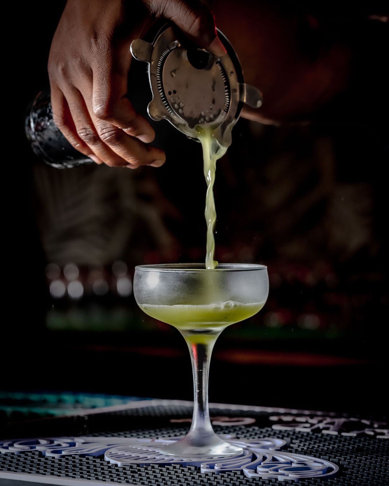 Bartender pouring a green cocktail through a strainer into a coupe glass on a dark bar.
