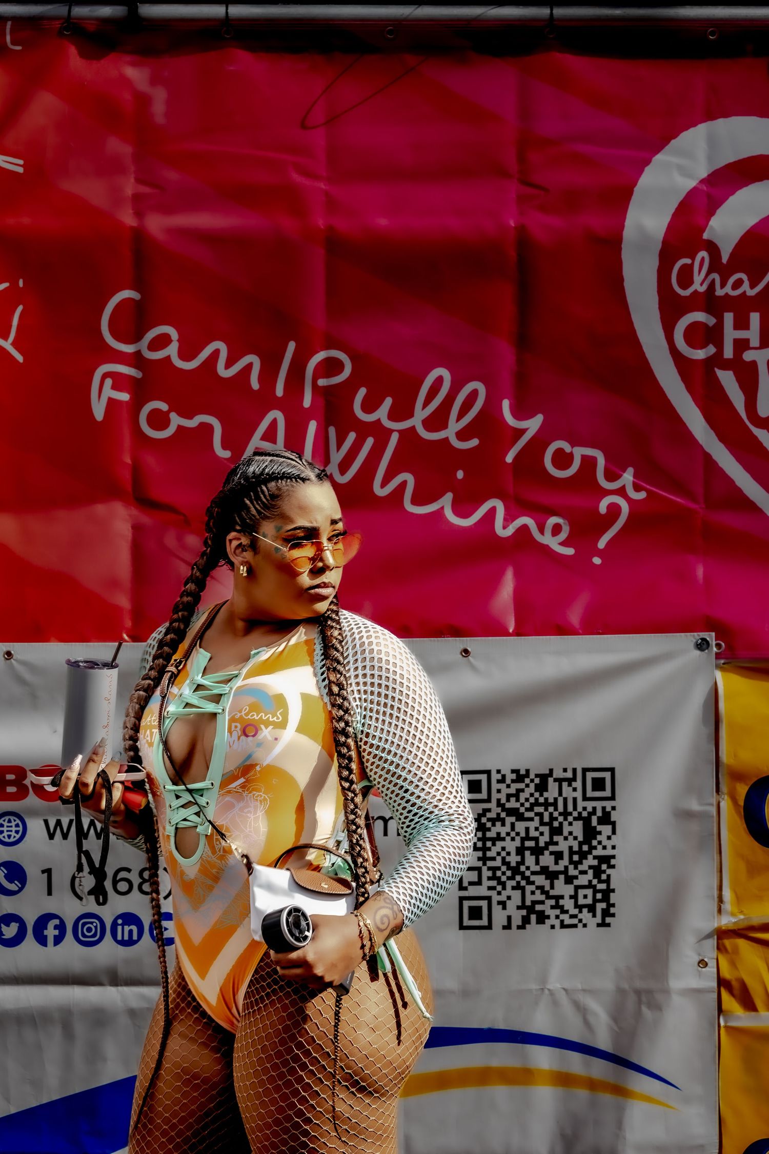 Woman in colorful outfit and braids poses in front of a red banner reading Can I Pull You For A Whine.