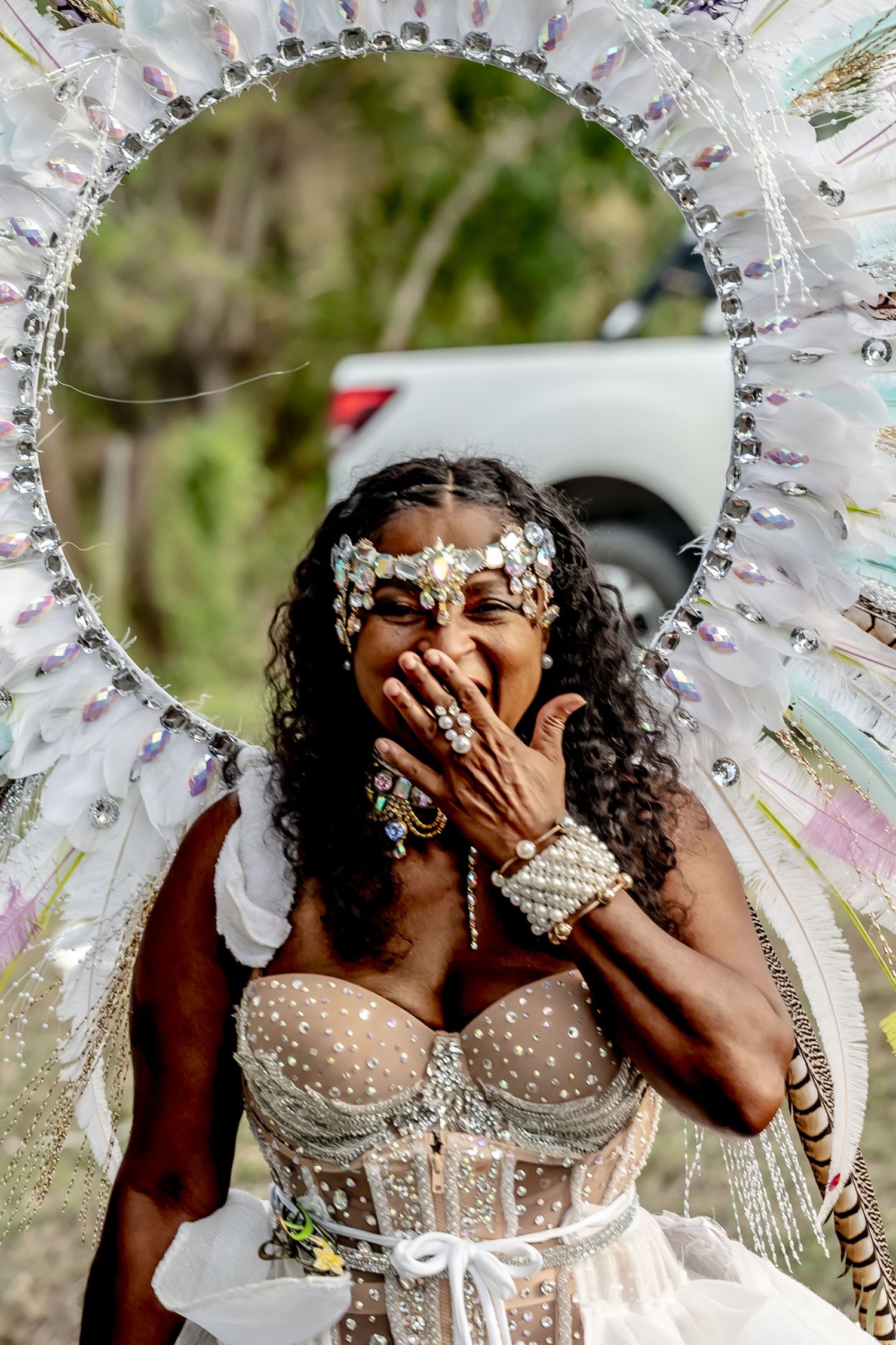 Woman in elaborate white carnival costume with feathered halo headdress and jeweled bodice laughing at outdoor festival.