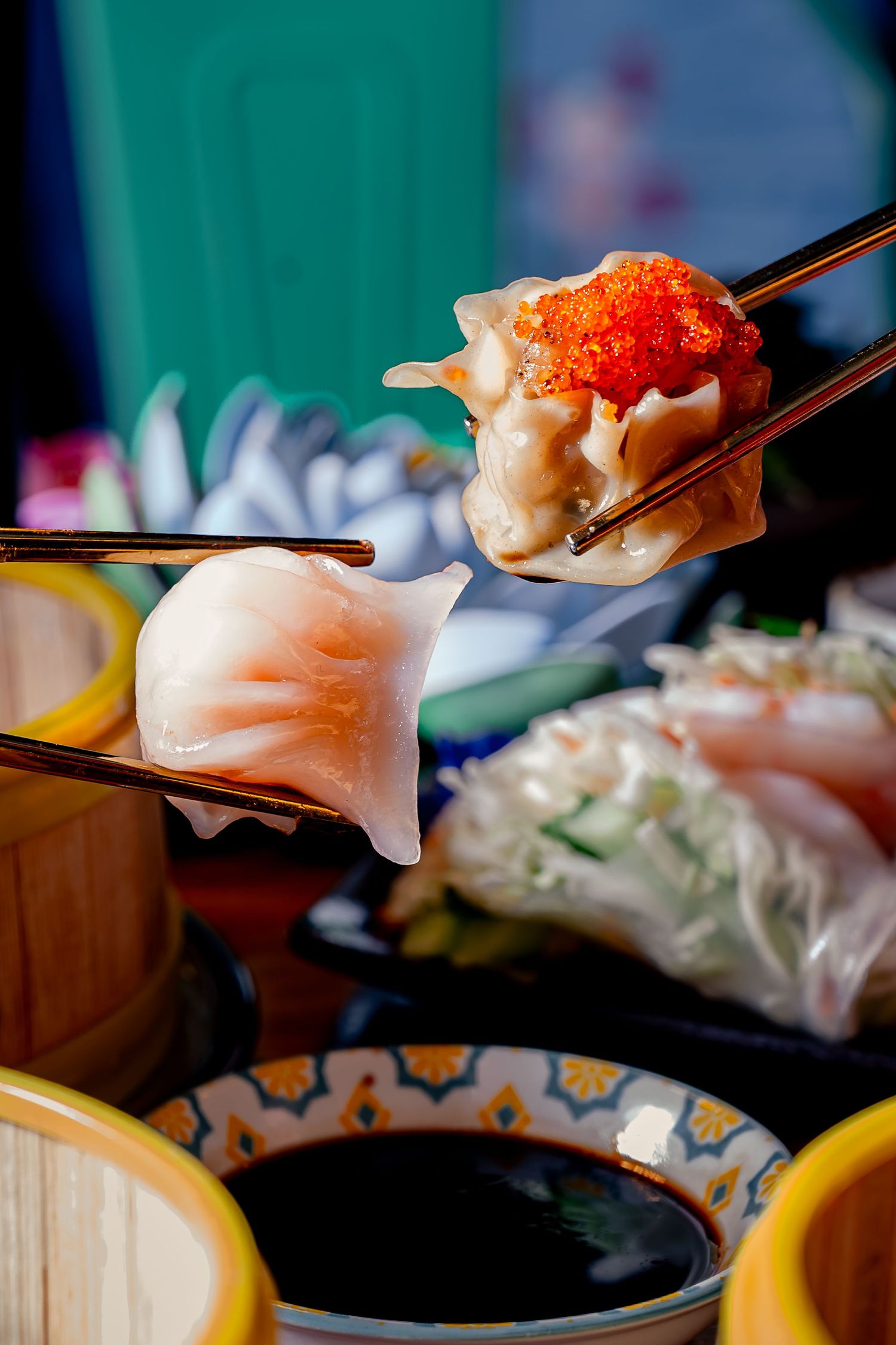 Chopsticks holding dumplings topped with fish roe over soy sauce, with dim sum and fresh rolls in background.