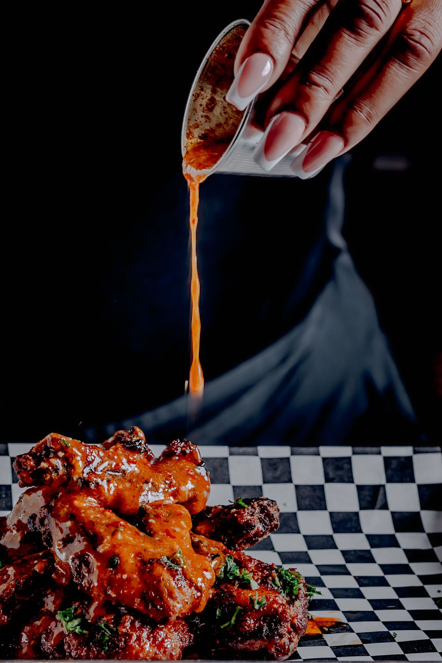 Hand pouring orange buffalo sauce over crispy chicken wings on checkered paper in dramatic dark lighting.