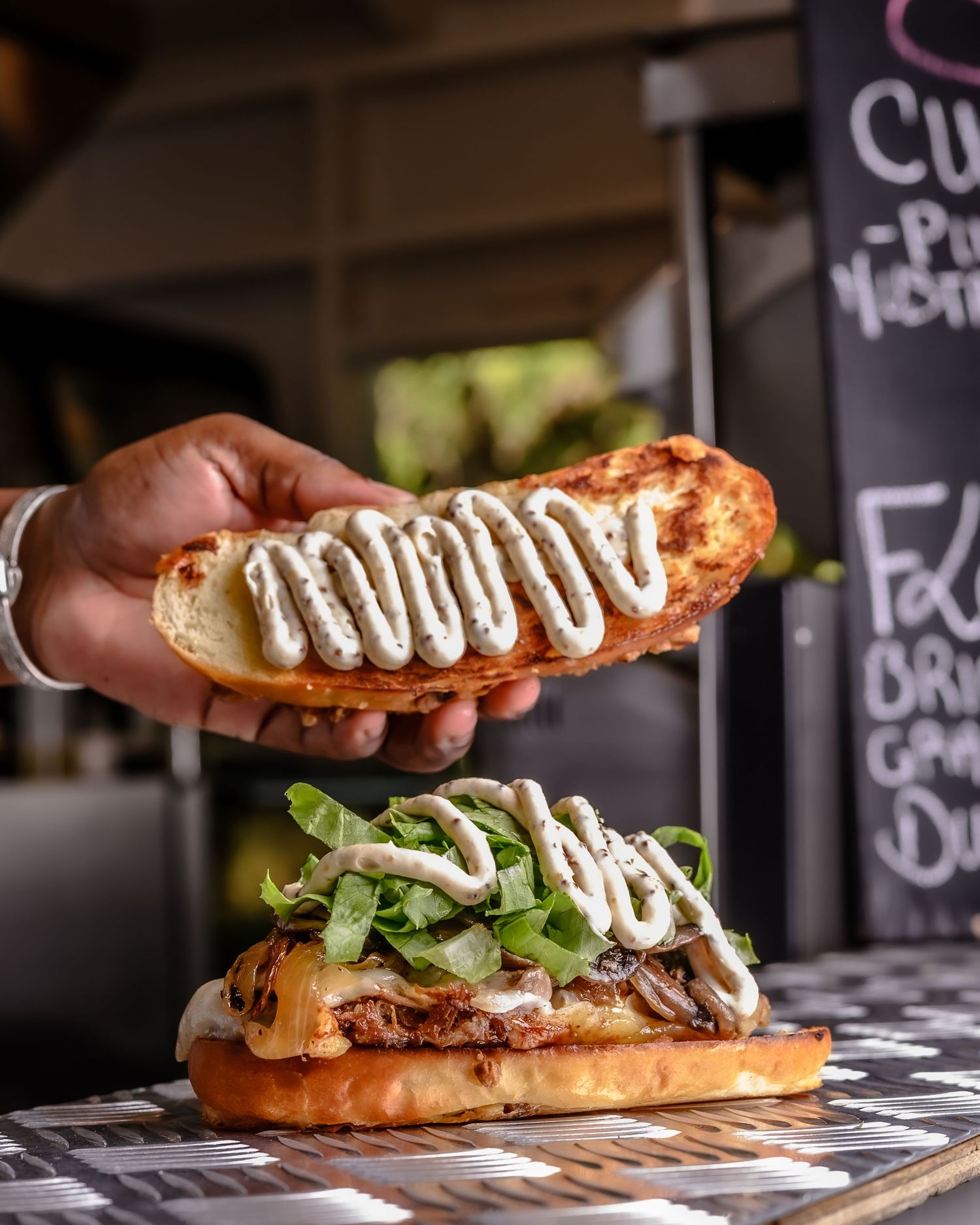 Hand holding a toasted bun drizzled with creamy sauce over a loaded burger with lettuce and toppings on a wooden board.