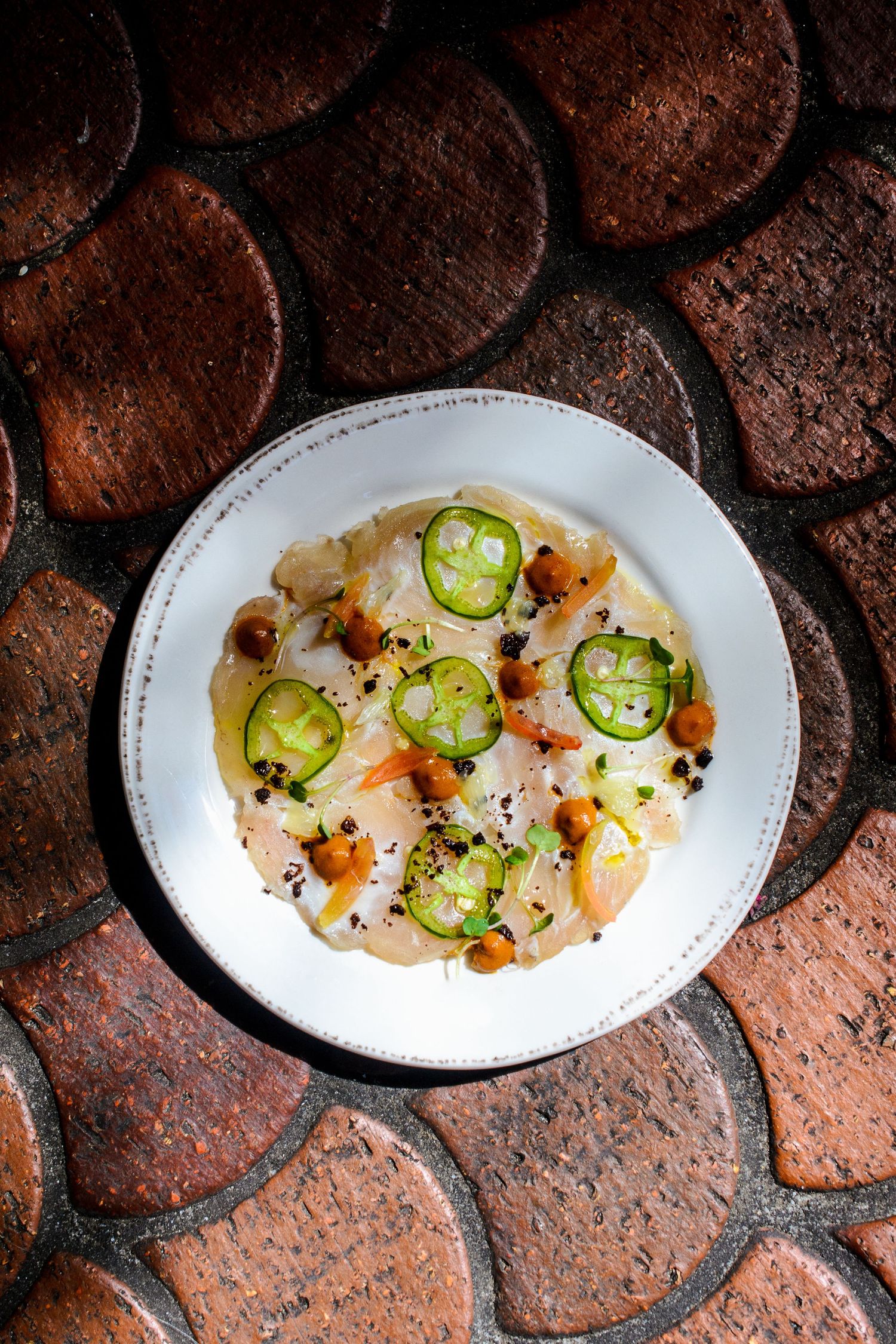 Elegant white plate with fish carpaccio, sliced jalapeños, chickpeas, and black sesame on rustic brick surface.