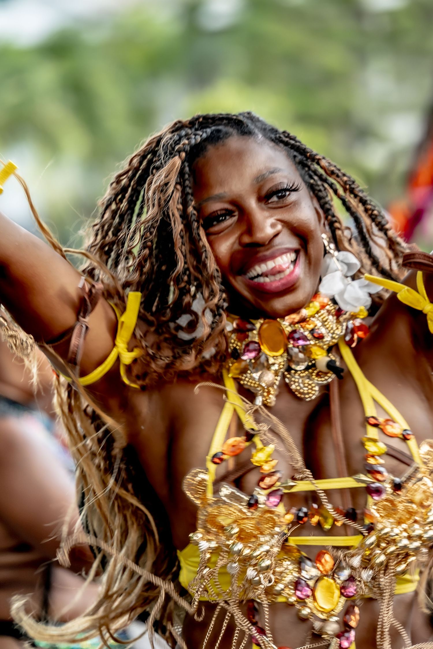 Smiling woman in golden carnival costume with braided hair and colorful beaded jewelry dancing at a festival.