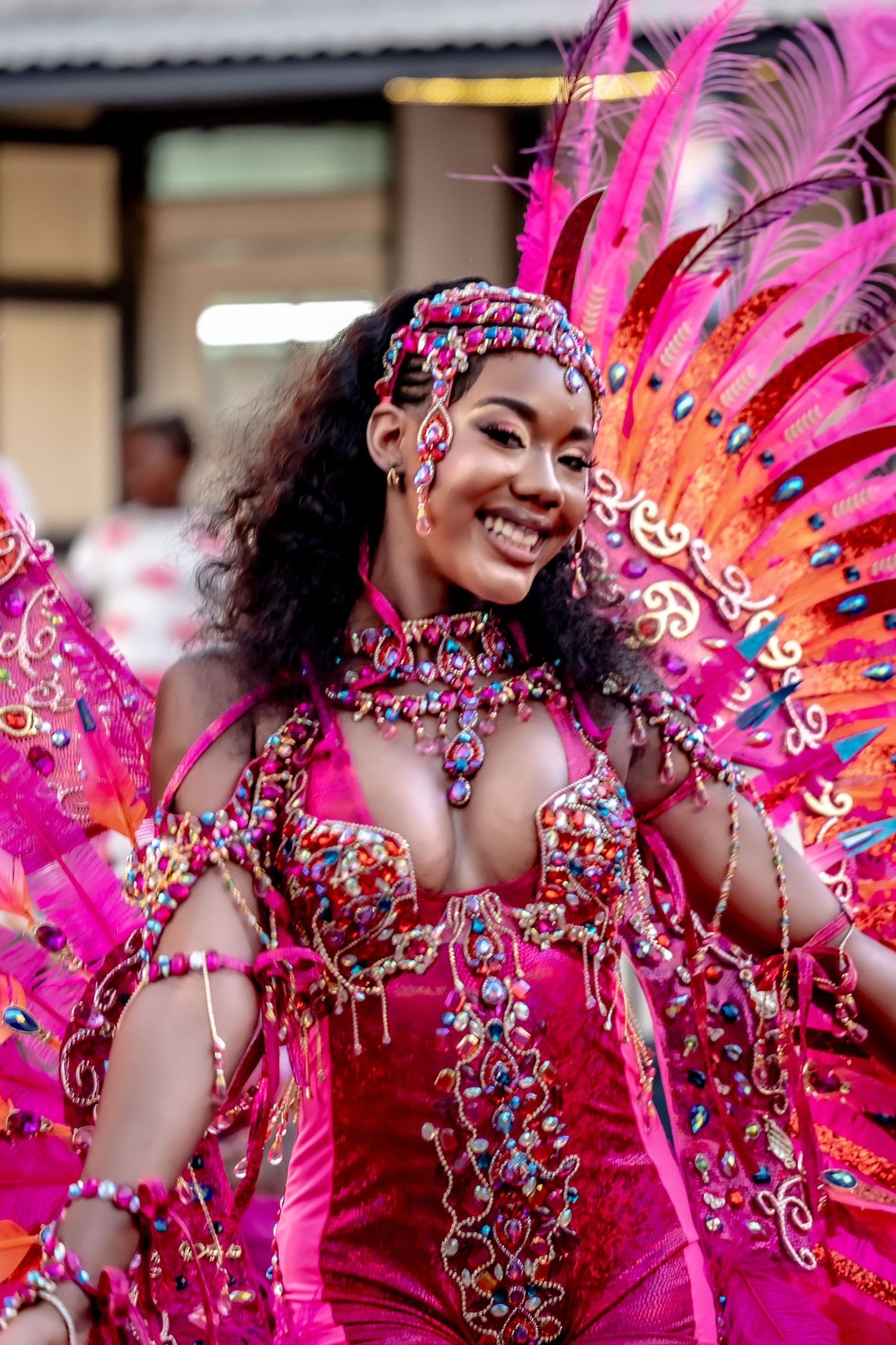 Smiling woman in vibrant pink carnival costume with feathers and jewels at a festive parade.