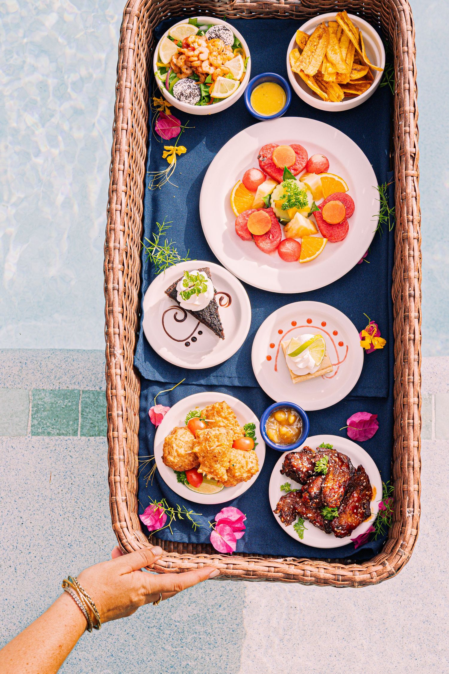 Overhead view of a wicker tray filled with colorful dishes, appetizers, and tropical foods held by a hand.