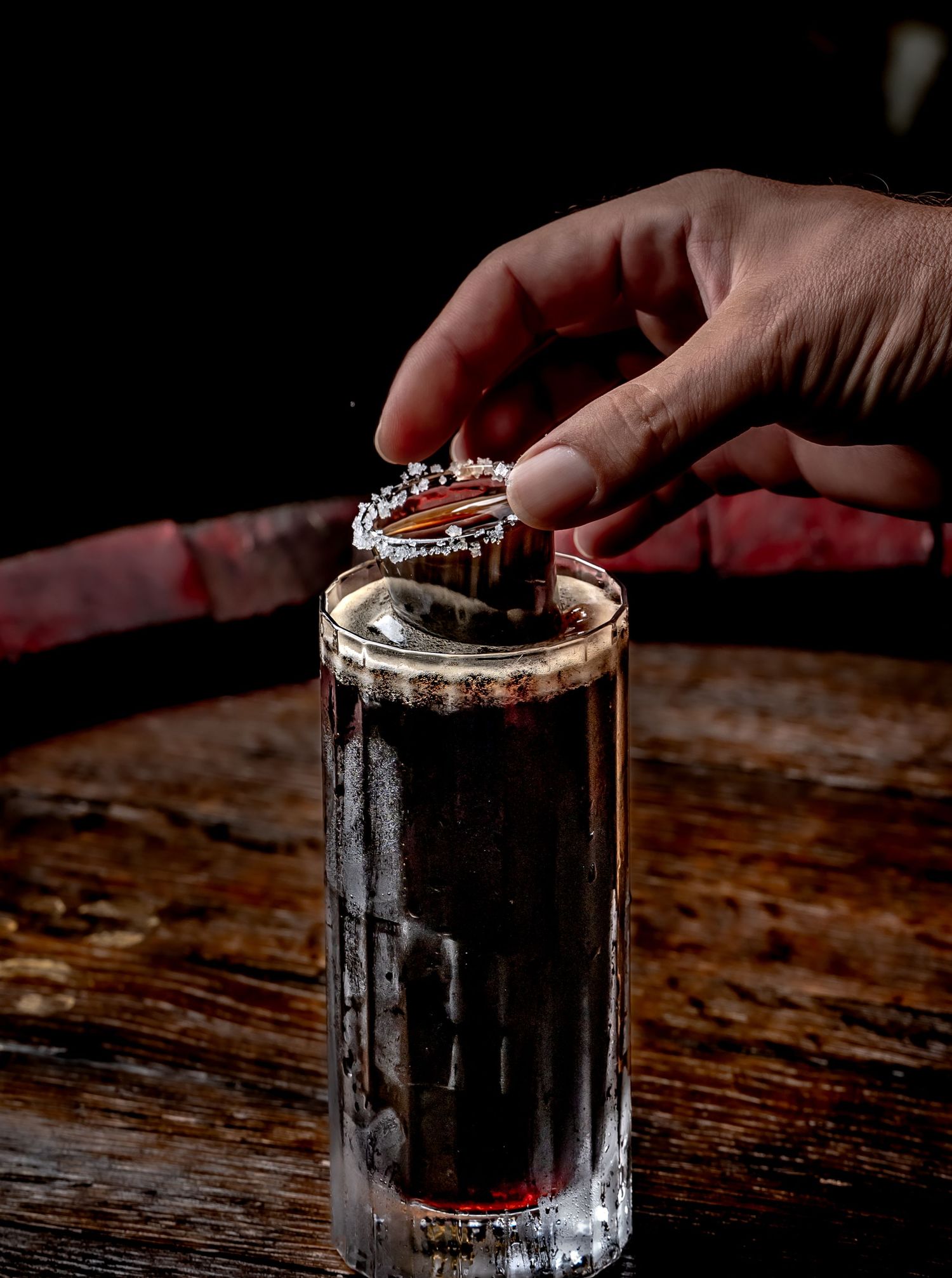 Hand placing a garnish on a dark cocktail shot glass on a rustic wooden bar.