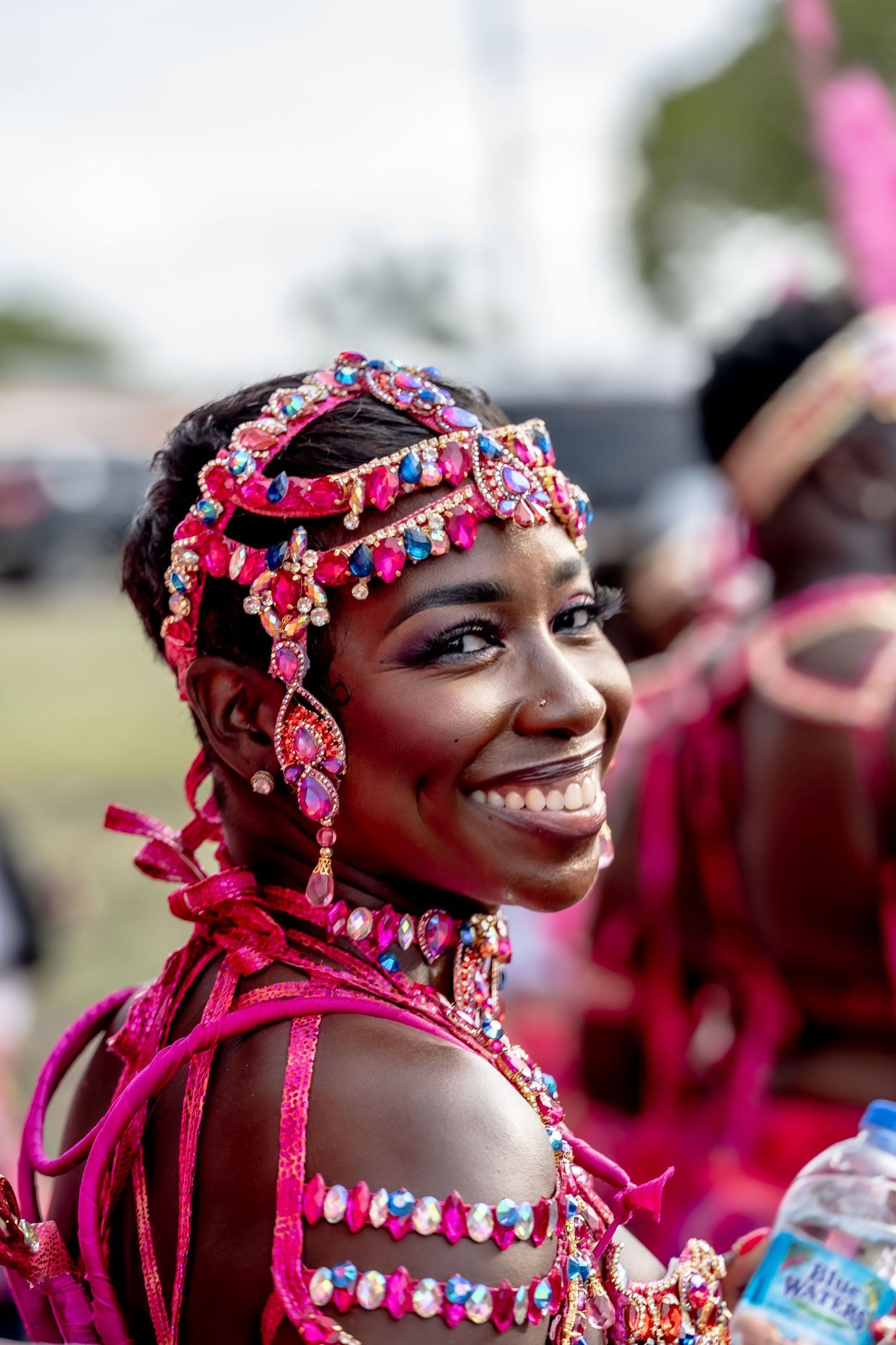 Smiling woman in vibrant pink jeweled carnival costume with ornate headpiece and matching accessories at a festival.