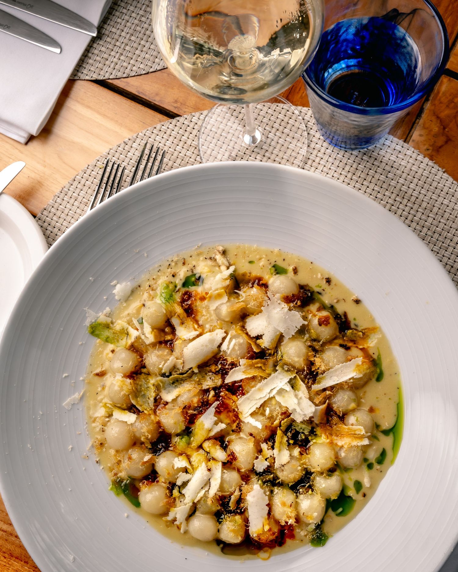 Elegant white bowl of gnocchi with crab, capers, and herbs in broth, served alongside white wine and blue water glass.