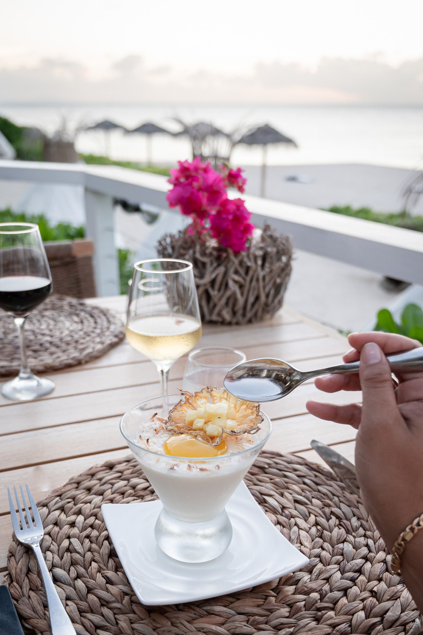 A dessert course with wine glasses and pink flowers on woven placemats at an outdoor beachfront restaurant setting.
