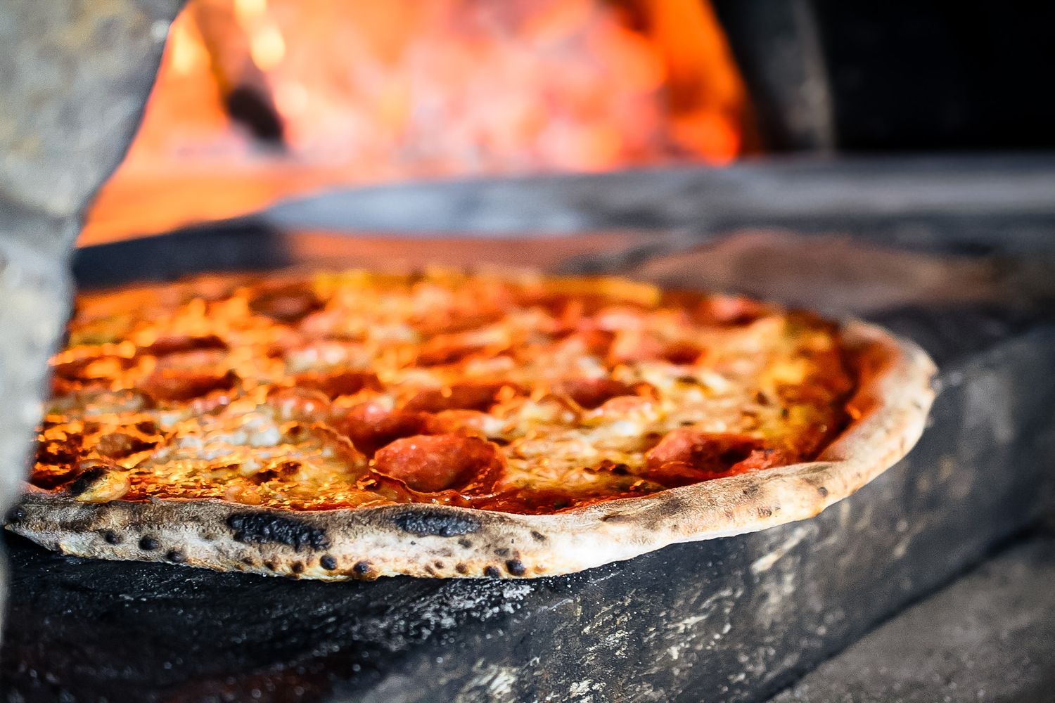 A wood-fired pizza bakes in a traditional brick oven with flames visible in the background.