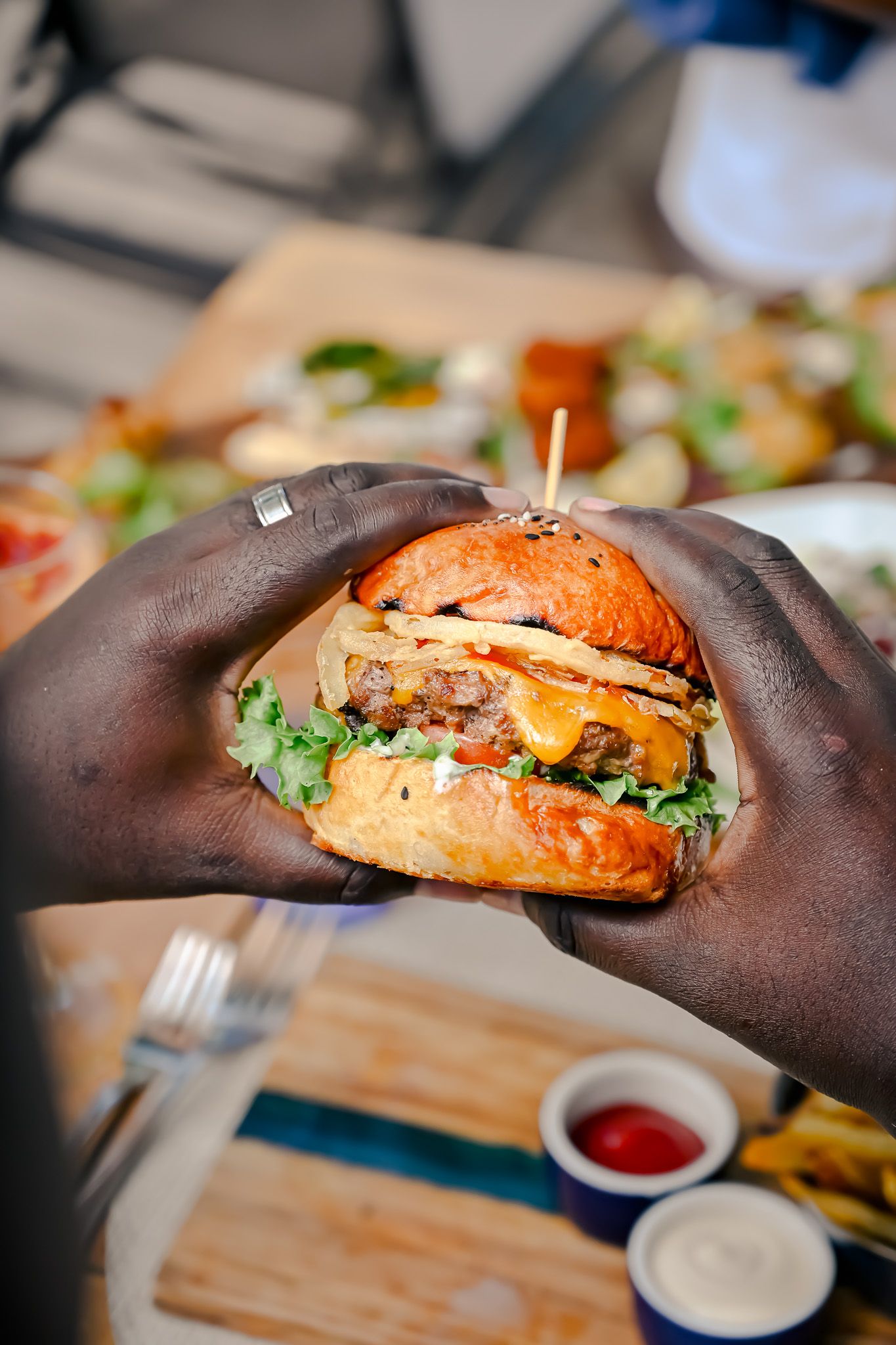 Hands holding a large double cheeseburger with lettuce on a wooden table with dipping sauces.