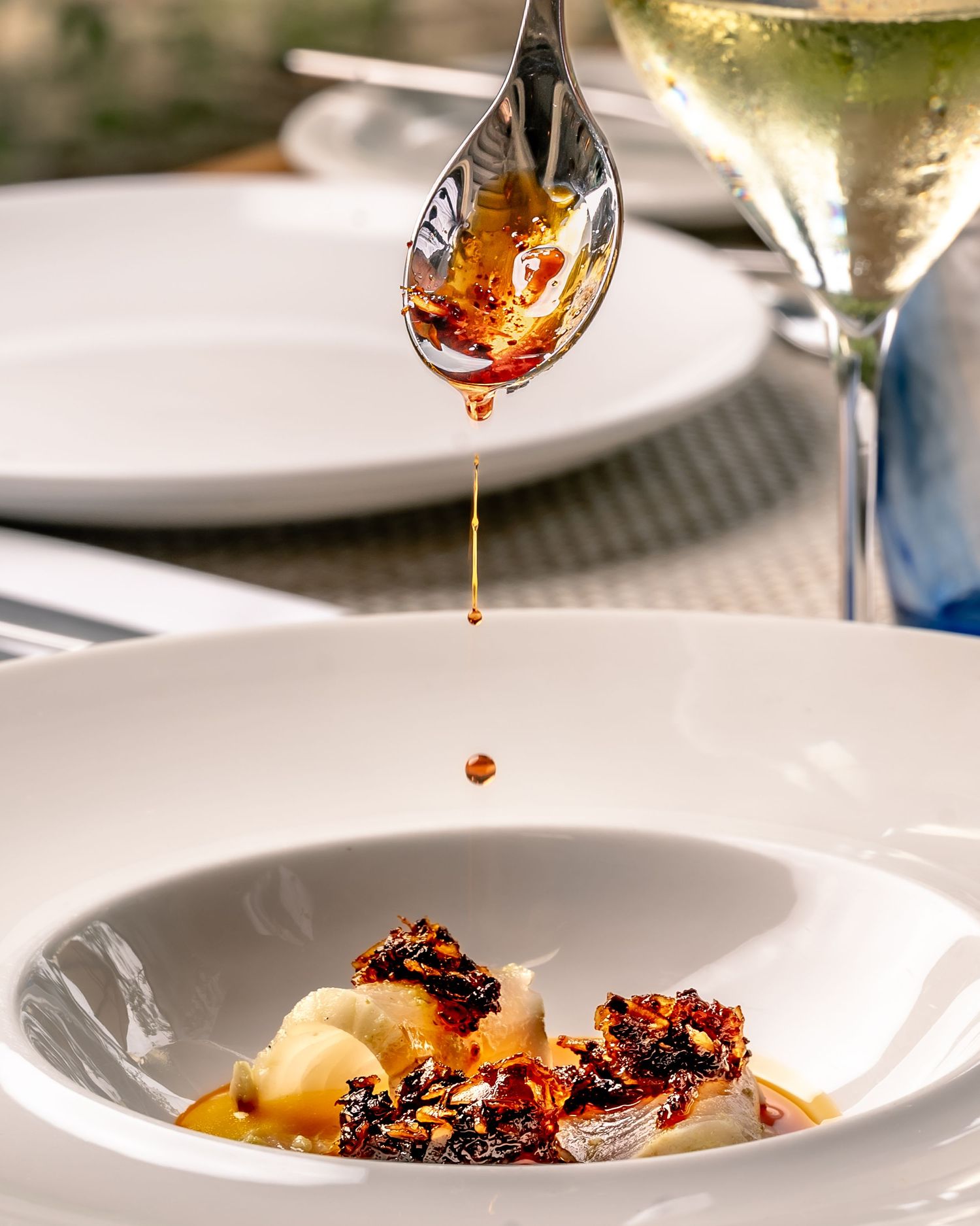 A spoon drizzling amber sauce over an elegant white bowl with gourmet dish, fine dining with wine glass in background.
